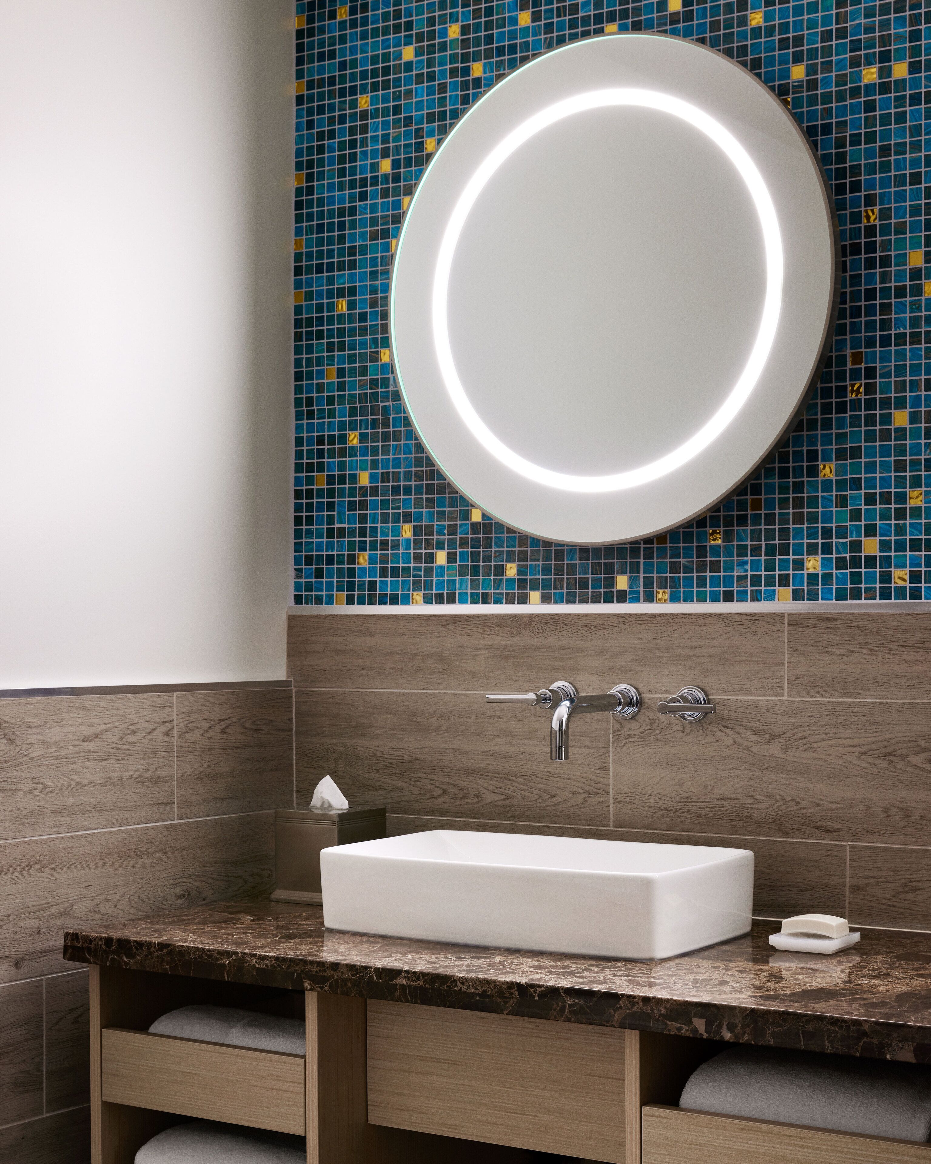 Un baño con un lavamanos blanco, un espejo redondo con luz y una pared de azulejos de mosaico azul.