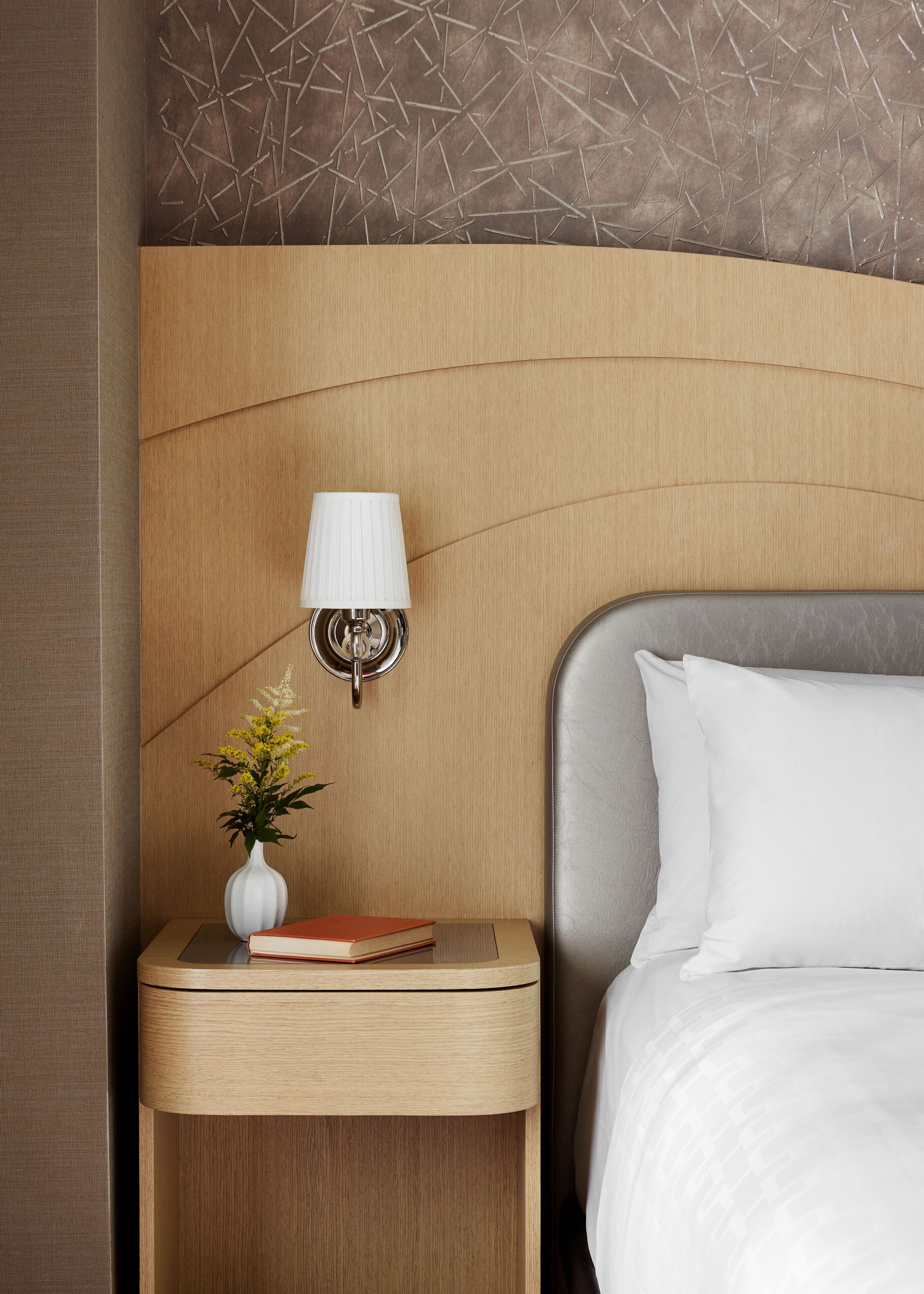 Habitación de hotel ordenada con cabecera de madera, lámpara de noche, mesita de noche con un jarrón de flores y un libro, y cama con ropa de cama blanca.