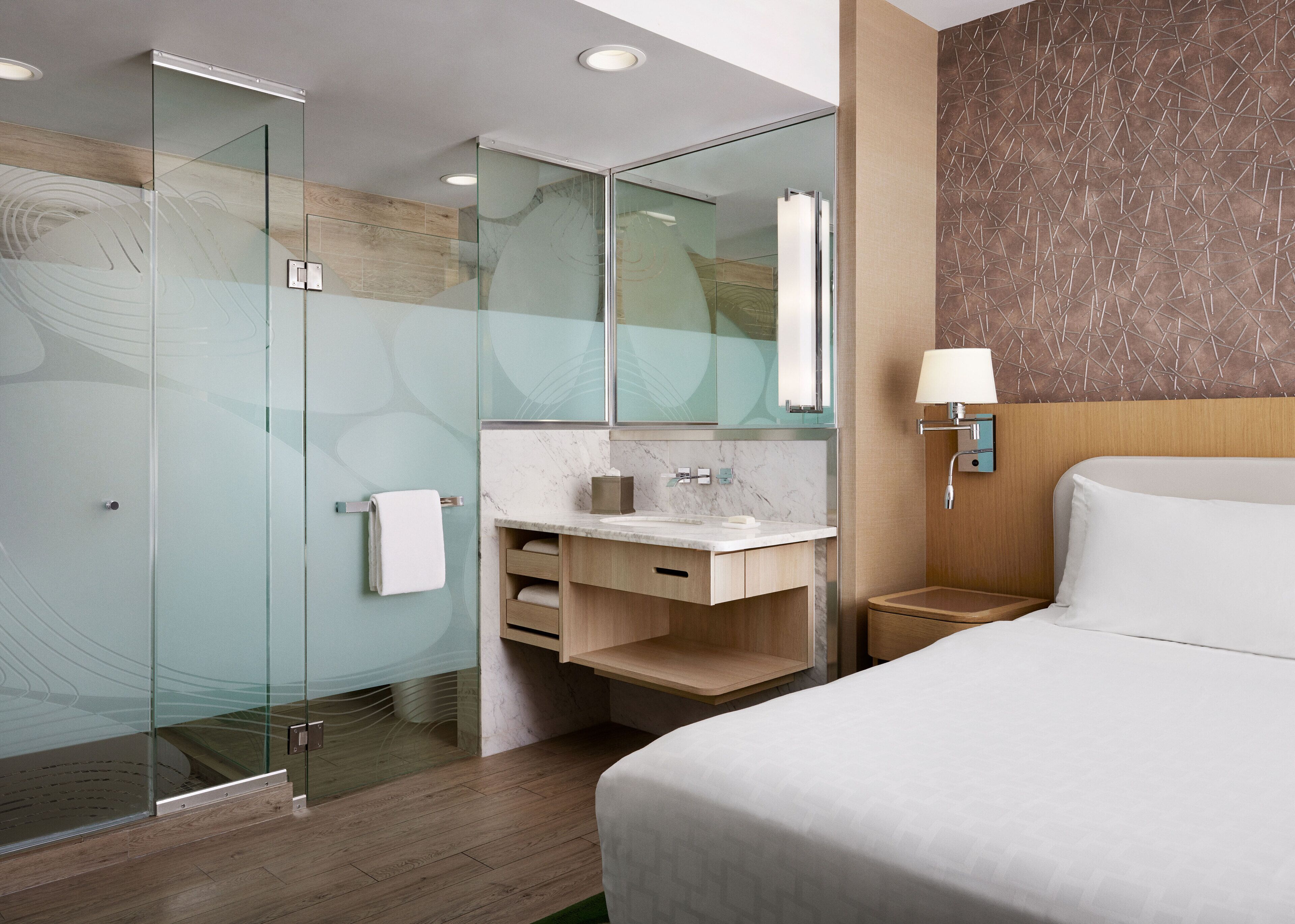 A modern hotel room with a glass-enclosed shower, a marble bathroom vanity, a wooden nightstand with a lamp, and a large bed with white linens.