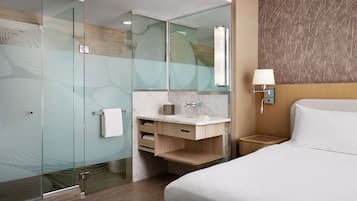 A modern hotel room with a glass-enclosed shower, a marble bathroom vanity, a wooden nightstand with a lamp, and a large bed with white linens.