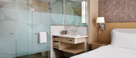 A modern hotel room with a glass-enclosed shower, a marble bathroom vanity, a wooden nightstand with a lamp, and a large bed with white linens.