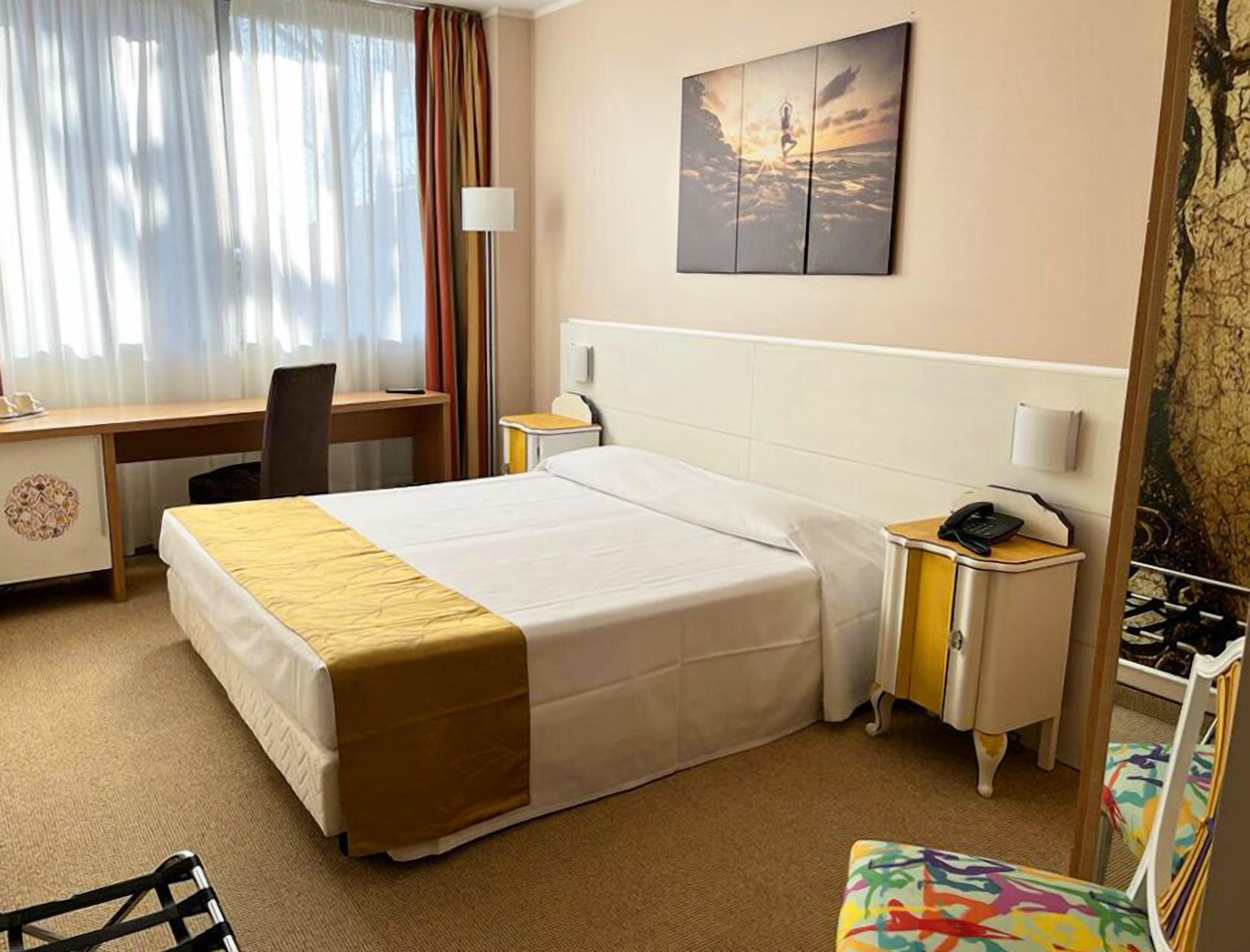 Habitación de hotel con cama, escritorio, silla, mesita de noche y un cuadro en la pared.