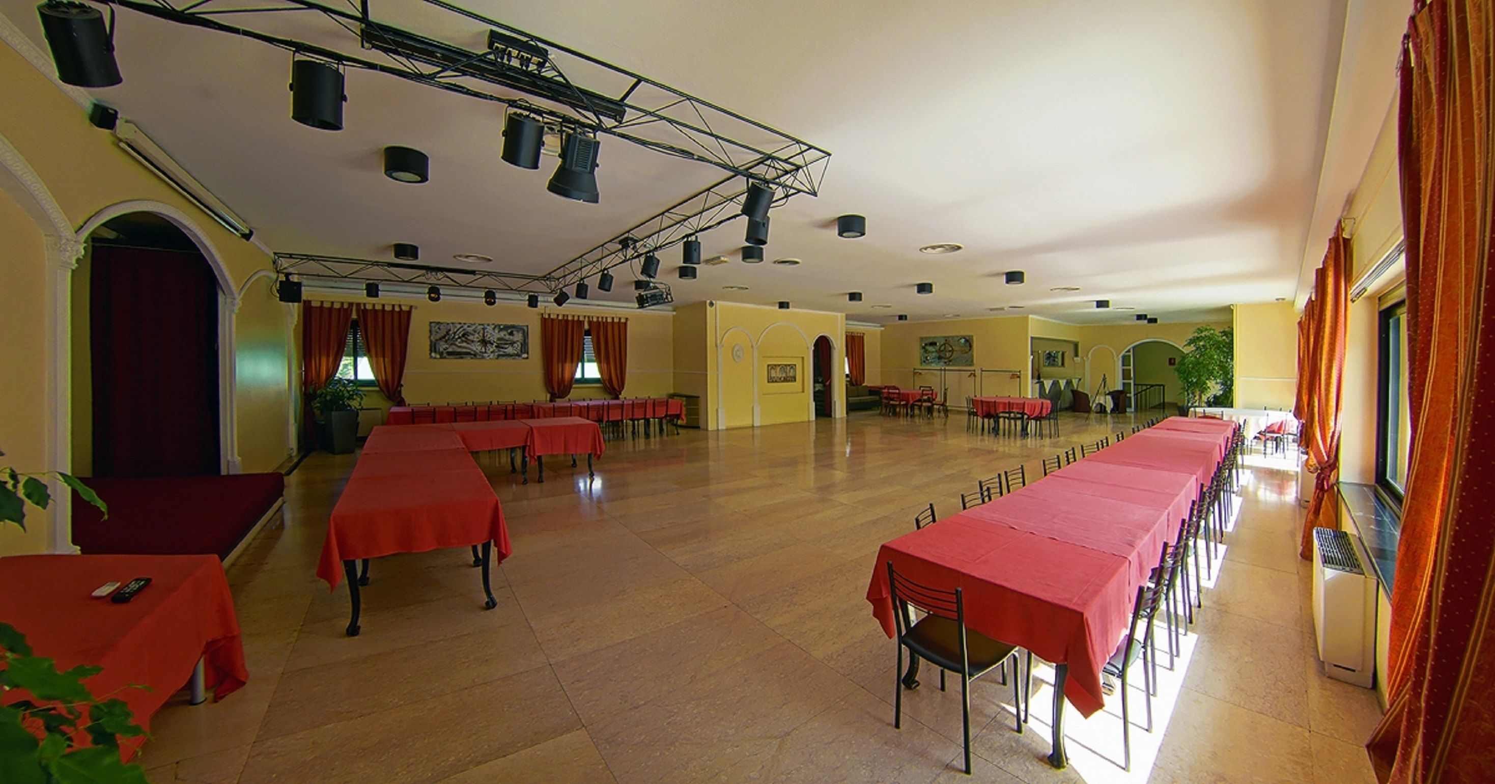 A large hall with rows of tables covered in red tablecloths, equipped with chairs, and a stage area with a red curtain.