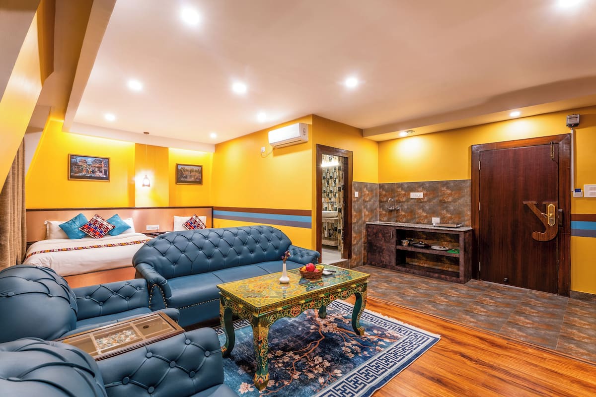 A hotel room with a blue sofa, a small table with a decorative item, a bed with colorful pillows, and a wooden cabinet.