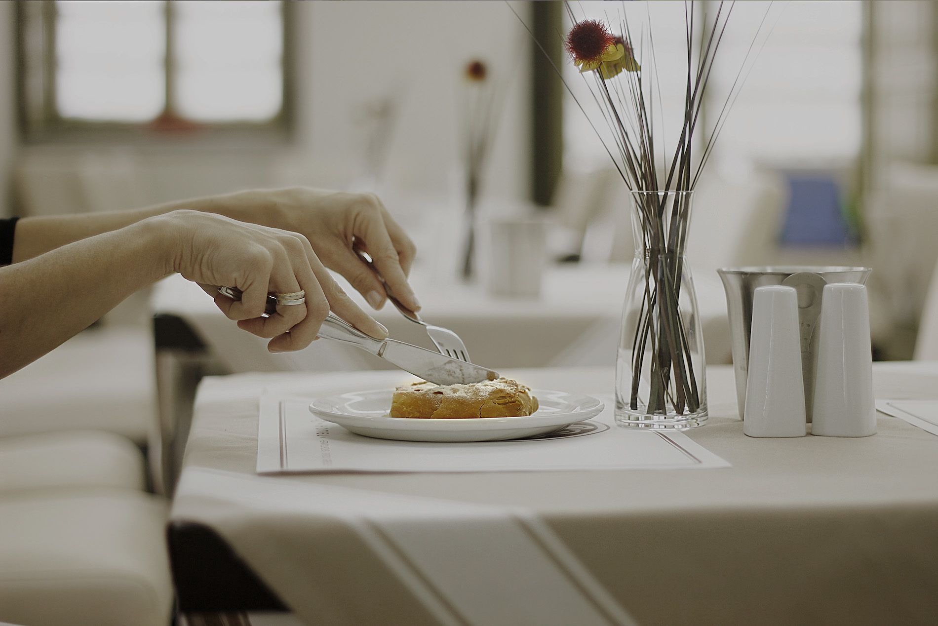 Una persona cortando un pastel sobre un plato blanco, en una mesa con un jarrón de flores secas y saleros.