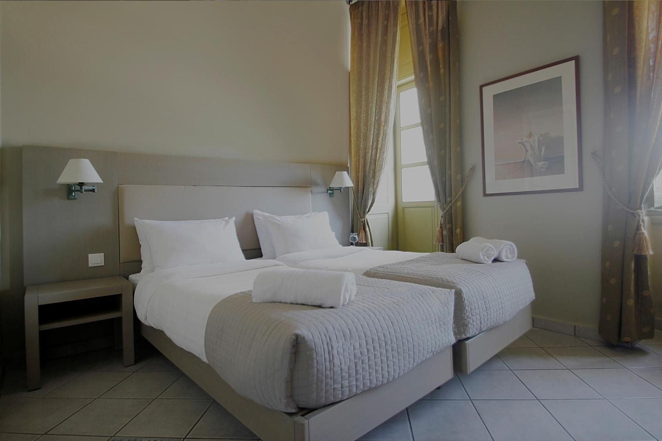 Una habitación de hotel con dos camas, una mesita de noche, una lámpara, un cuadro en la pared y cortinas.