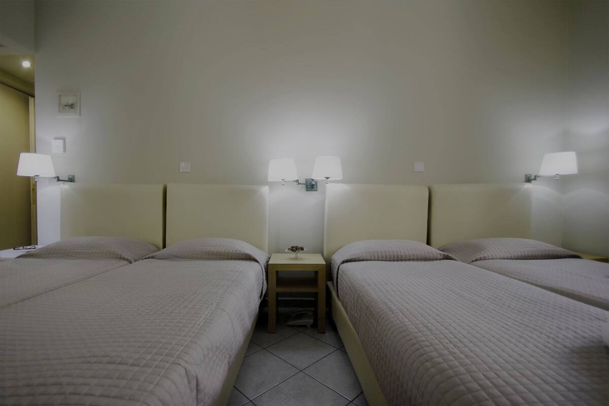 Quarto de hotel com duas camas de solteiro, uma mesinha de cabeceira com abajur e uma pequena mesa com um vaso.