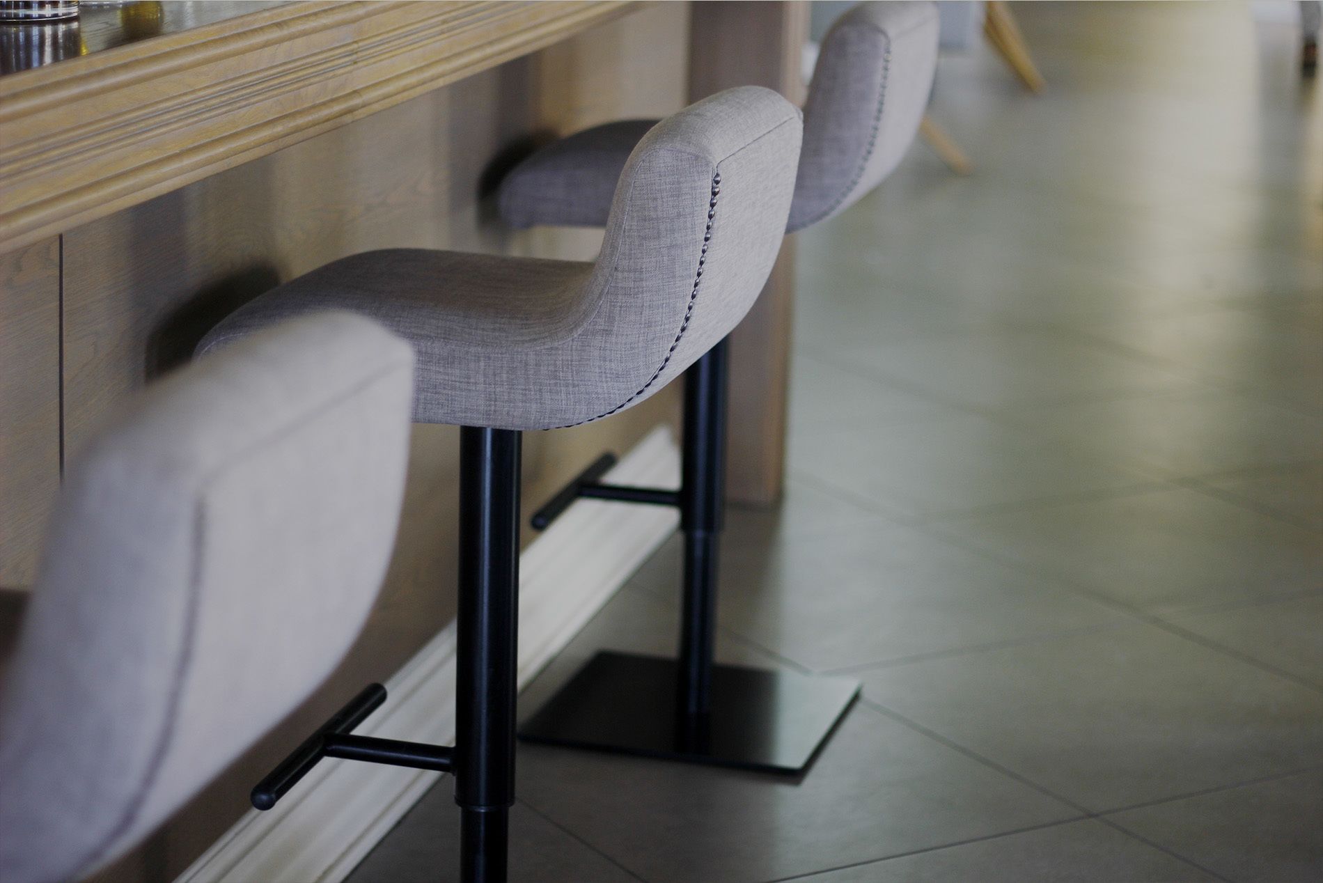 Una hilera de taburetes de bar con asientos tapizados en gris y patas de metal negro, colocados frente a una pared con mostrador de madera.
