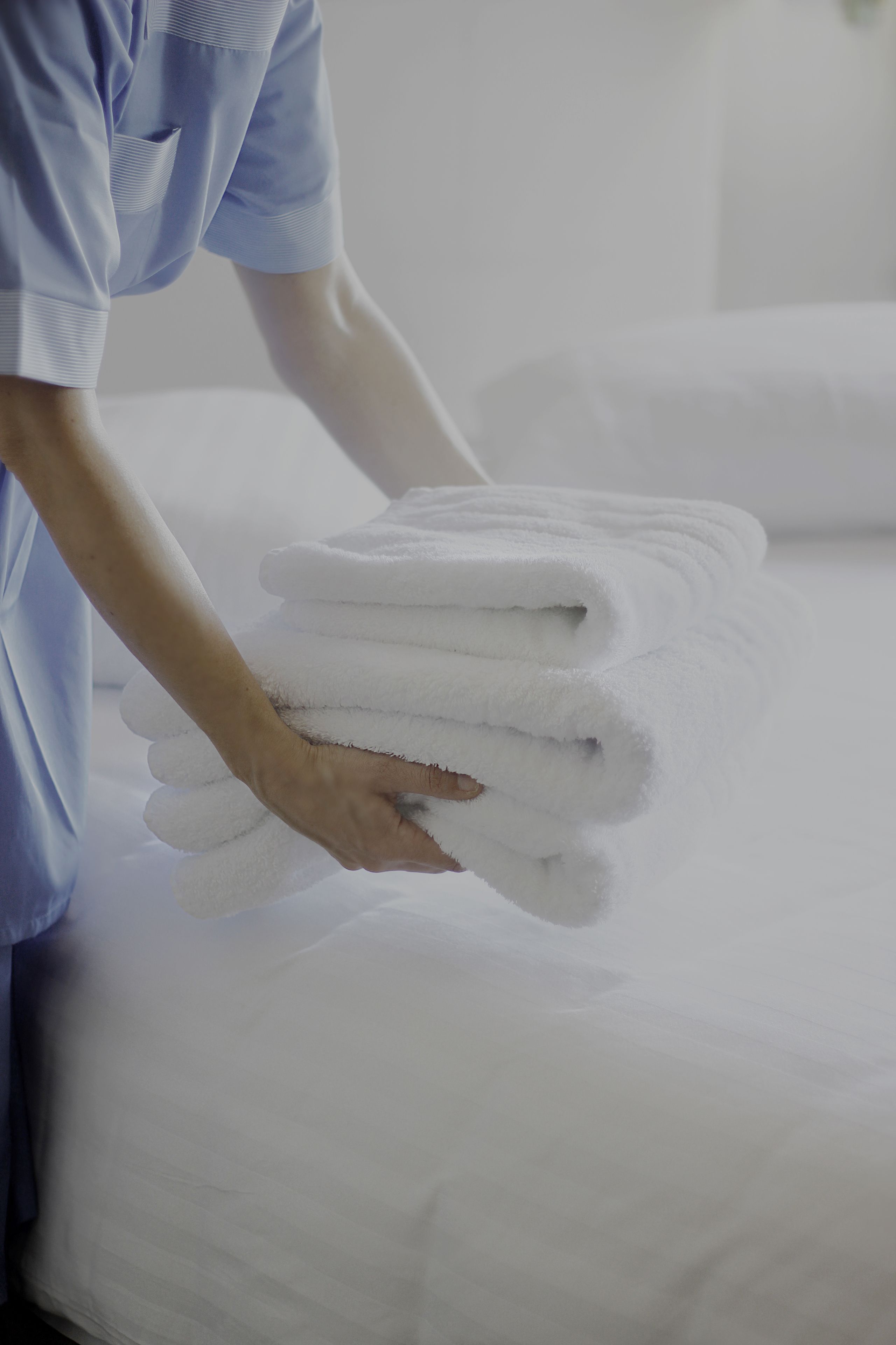 A person holding a stack of white towels on a bed with white sheets.