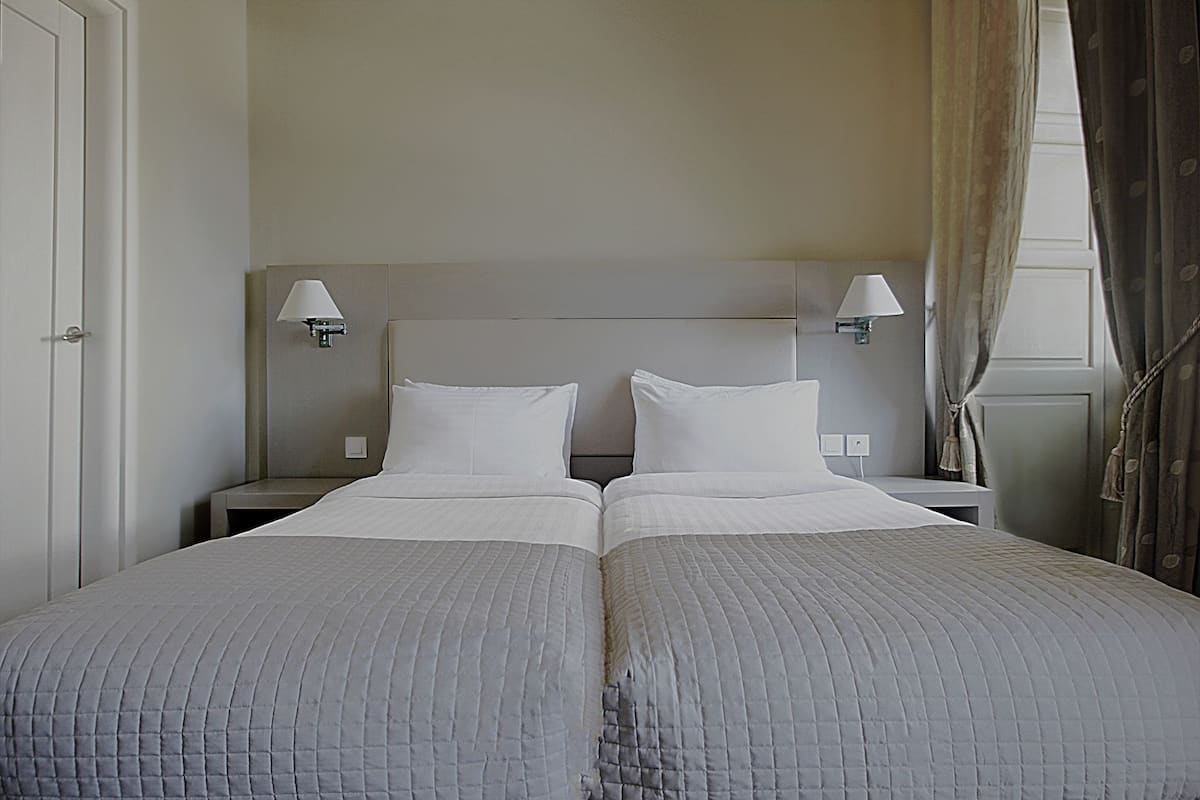 Cama de casal com duas almofadas e cabeceira, ladeada por mesas de cabeceira com abajures, em um quarto de tons neutros.