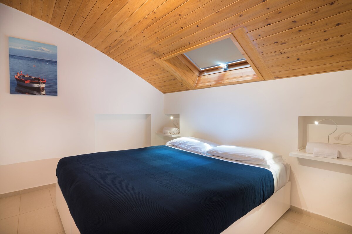 A bedroom with a wooden ceiling, a skylight, a bed with white and blue bedding, a painting of a boat, and a small shelf with a lamp.