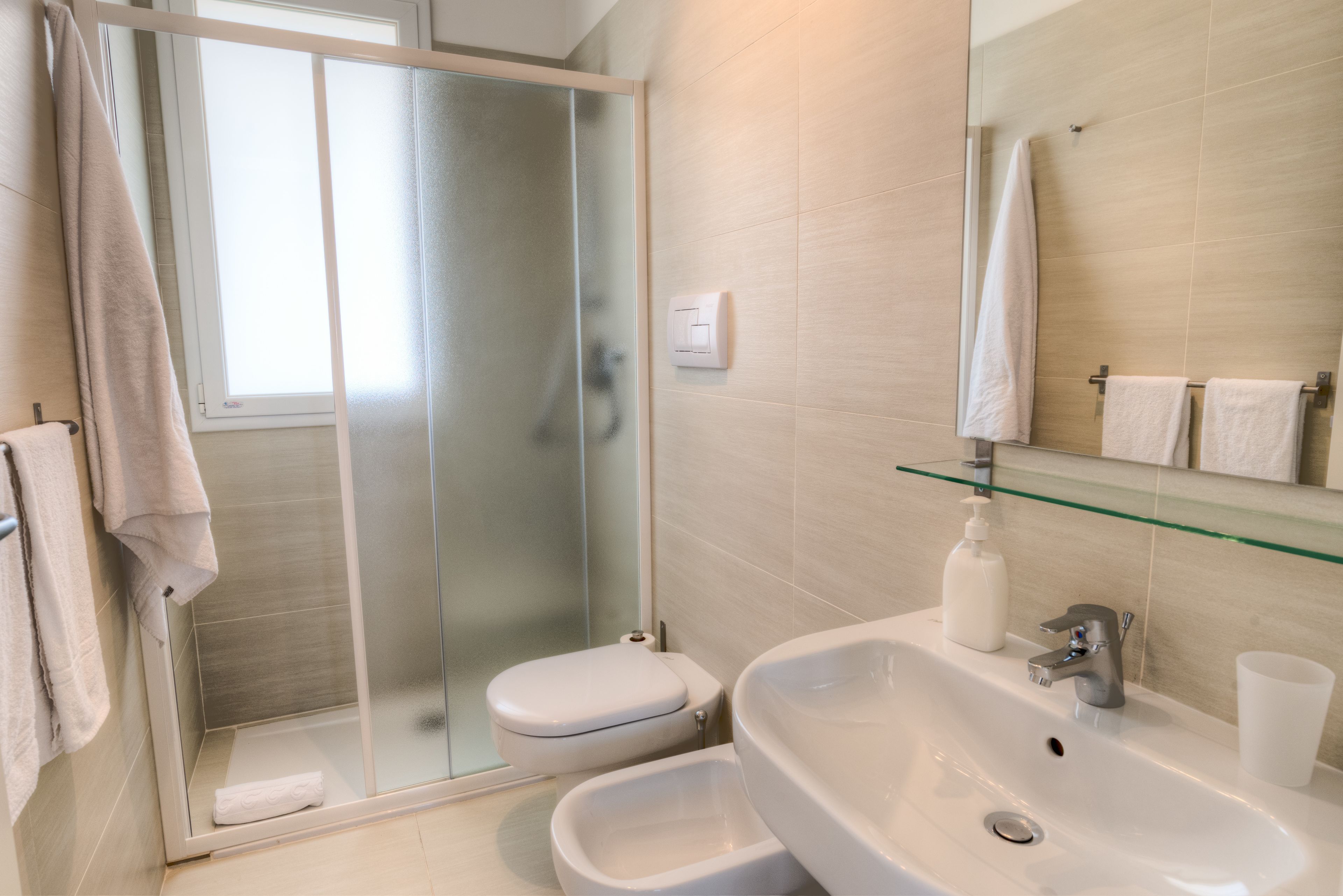 A modern bathroom with a glass shower enclosure, a toilet, a sink with a soap dispenser, and a glass shelf.