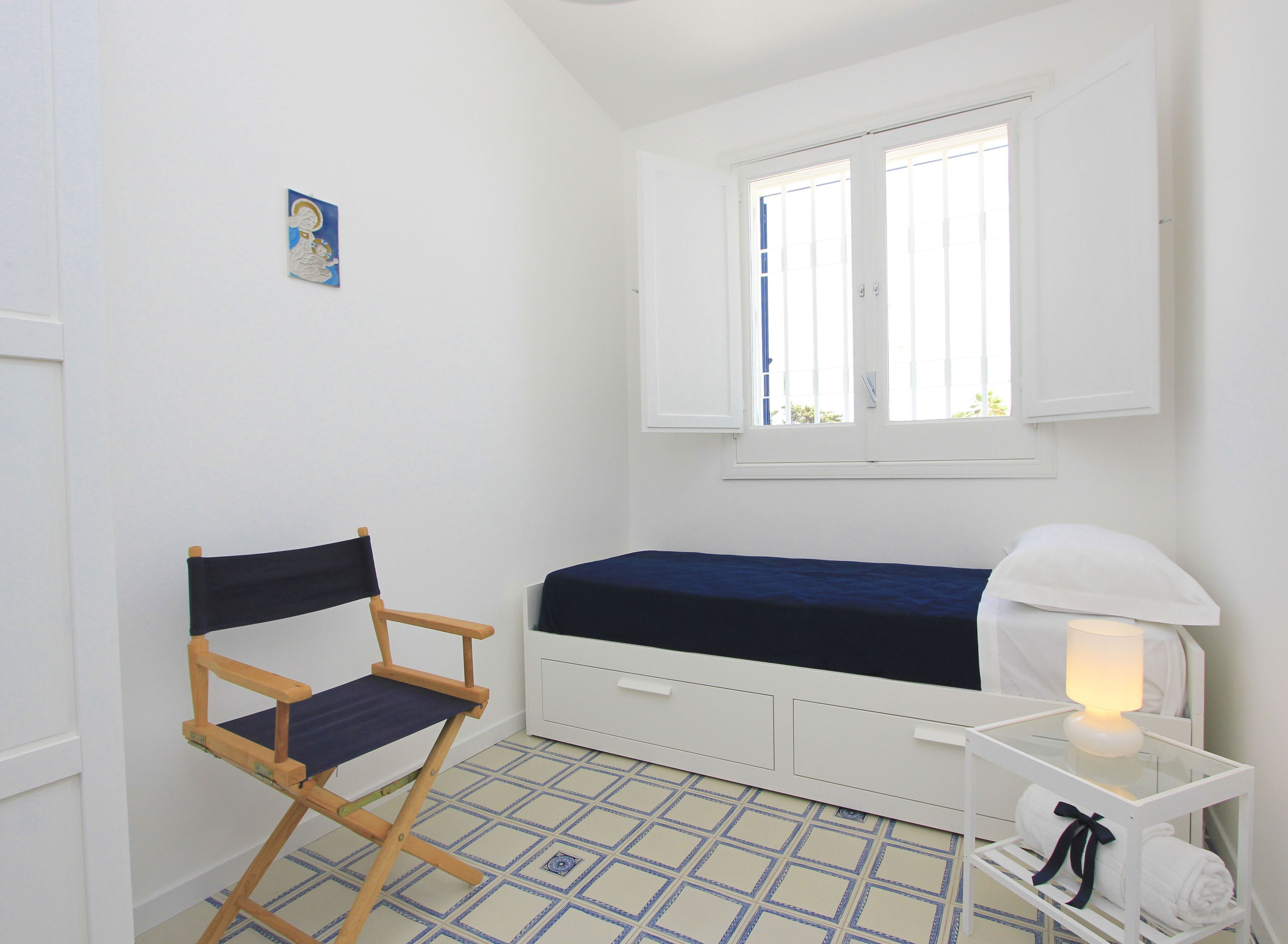 Een kleine, lichte kamer met een enkel bed, een opklapbare stoel, een nachtkastje met een lamp en een raam met zonwering.