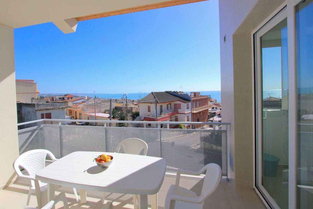 A balcony with a table and chairs, a bowl of fruit, and a view of the sea and buildings.