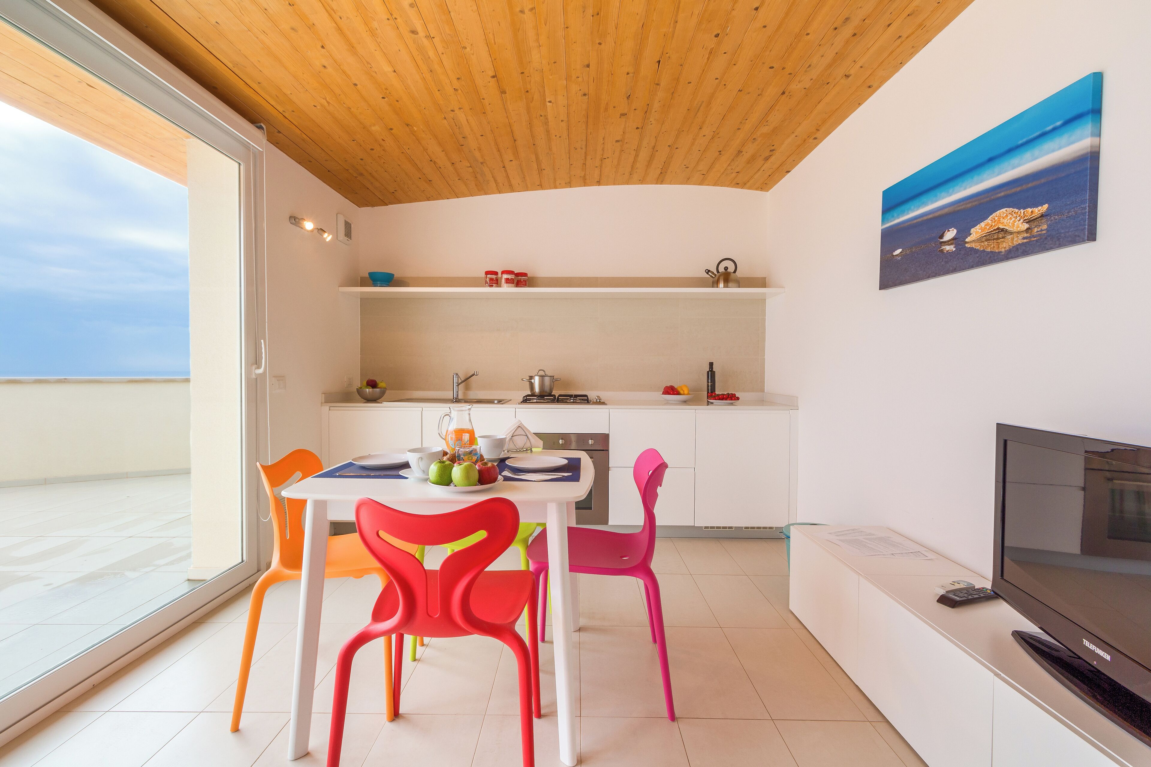 Una cocina moderna con armarios blancos, una zona de comedor con sillas coloridas y un ventanal con vistas a un balcón.