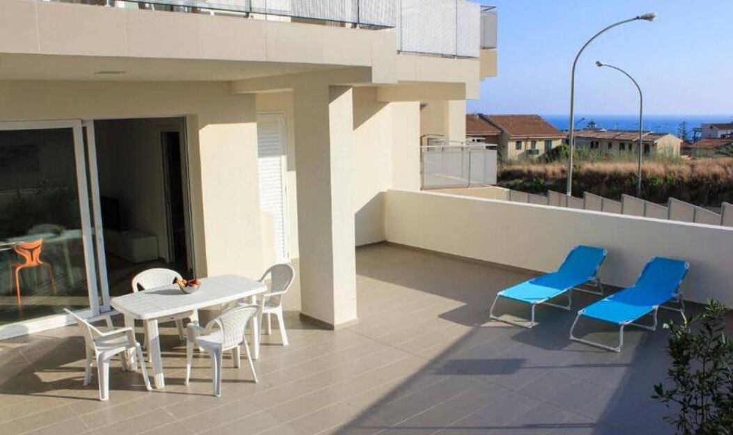 Eine moderne Außenpatio mit Tisch und Stühlen, eine Sitzecke mit blauen Liegestühlen und Blick auf das Meer und Gebäude im Hintergrund.