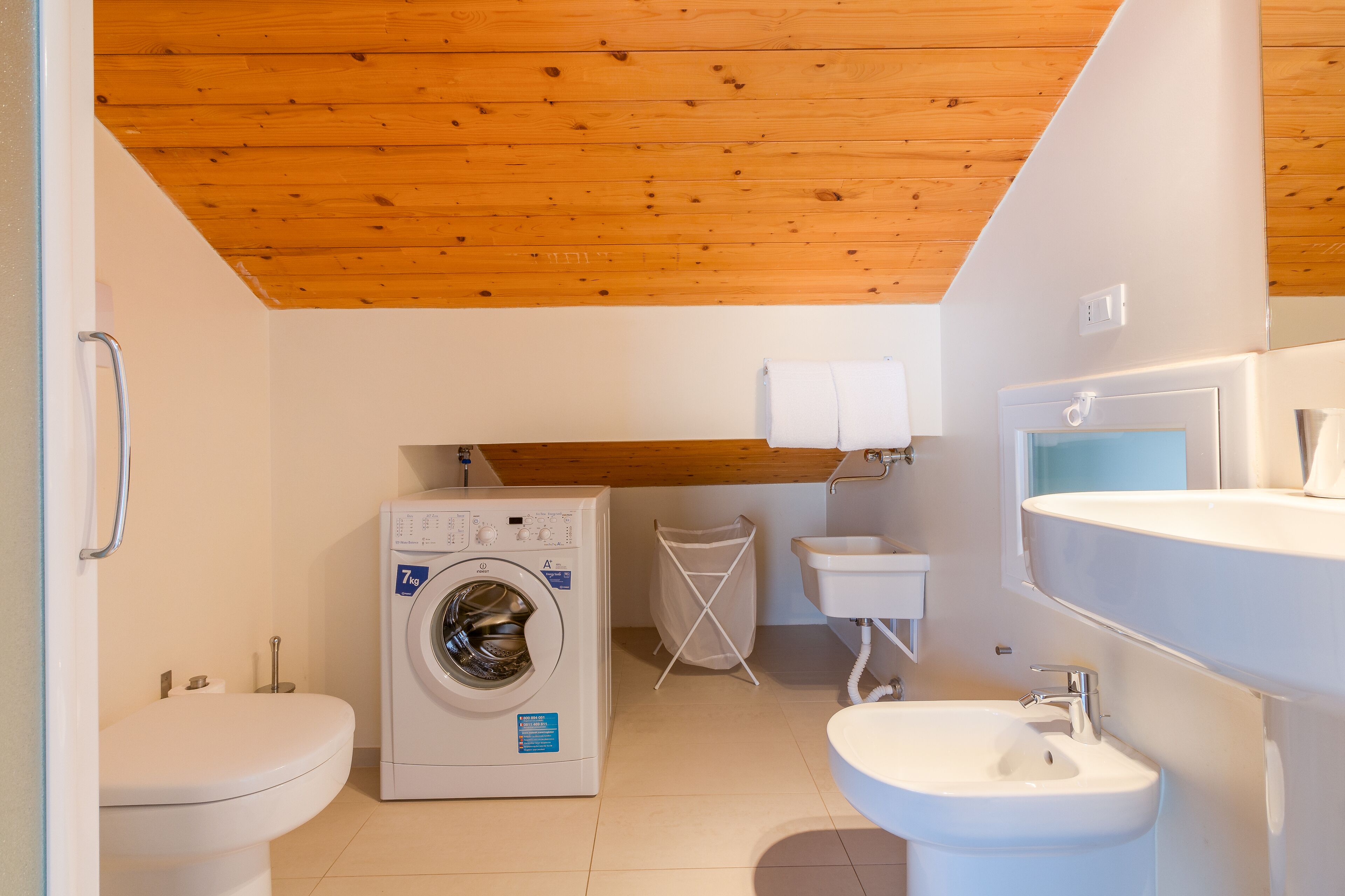 A compact bathroom with a washing machine, toilet, sink, and a wooden ceiling.
