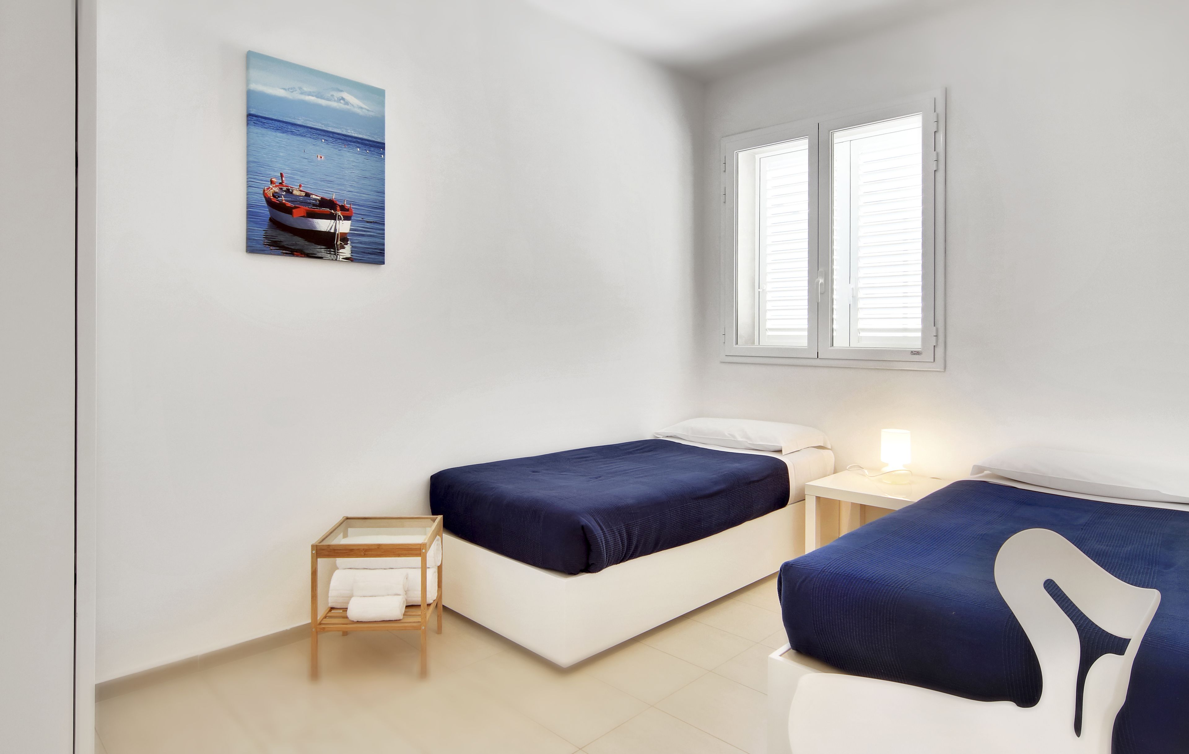 A bedroom with two beds, a small bedside table, a nightstand, a window with blinds, and a painting of a boat on the wall.