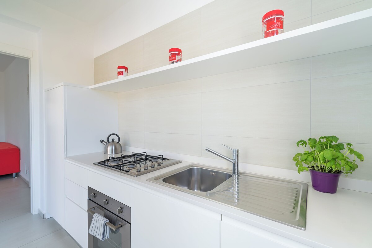 A modern kitchen with a stainless steel sink, a gas stove, and a kettle. There are white cabinets and a red chair in the background.