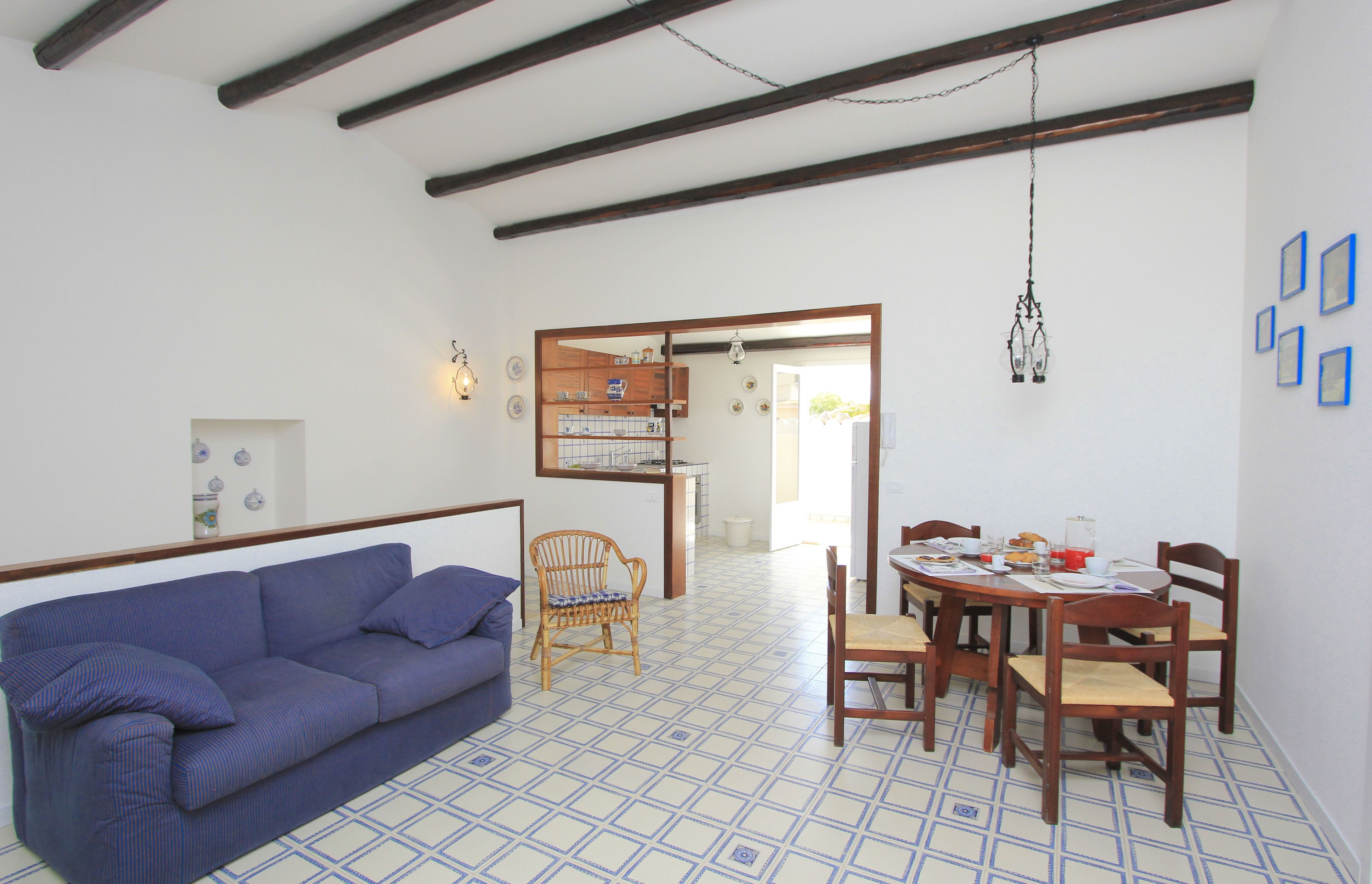 Una habitación con un sofá azul, una mesa de comedor de madera y sillas, una silla de mimbre y suelo de baldosas.