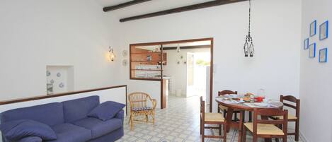 Una habitación con un sofá azul, una mesa de comedor de madera y sillas, una silla de mimbre y suelo de baldosas.