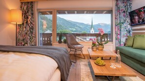 Une chambre d’hôtel avec un lit, une table basse en bois sur laquelle sont disposés des fruits et des boissons, un canapé vert, un fauteuil et une grande fenêtre donnant sur une église et des montagnes.