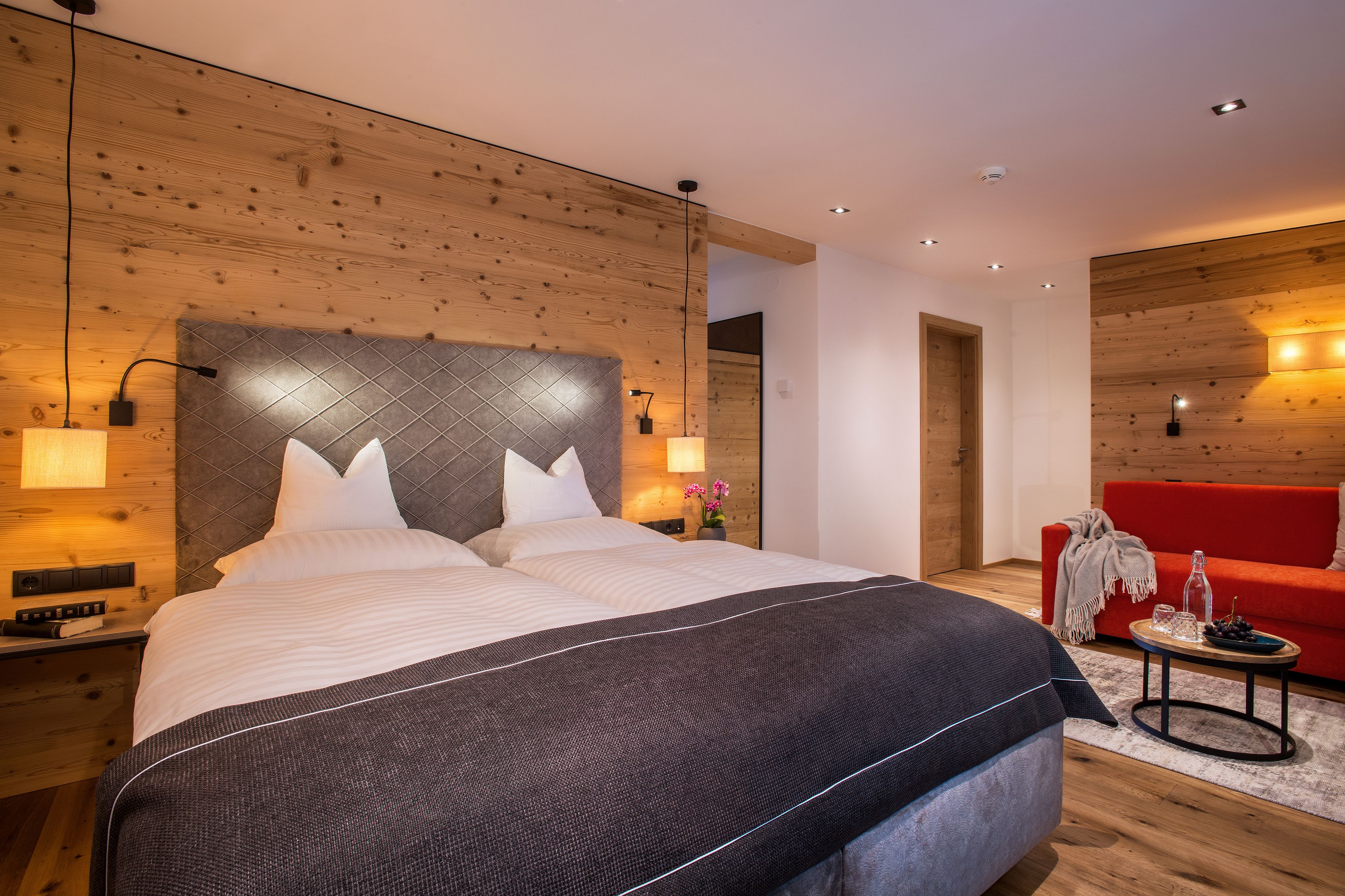 Un dormitorio moderno con una cama grande, cabecera de madera, lámparas de noche, un sofá rojo y una mesita con adornos.