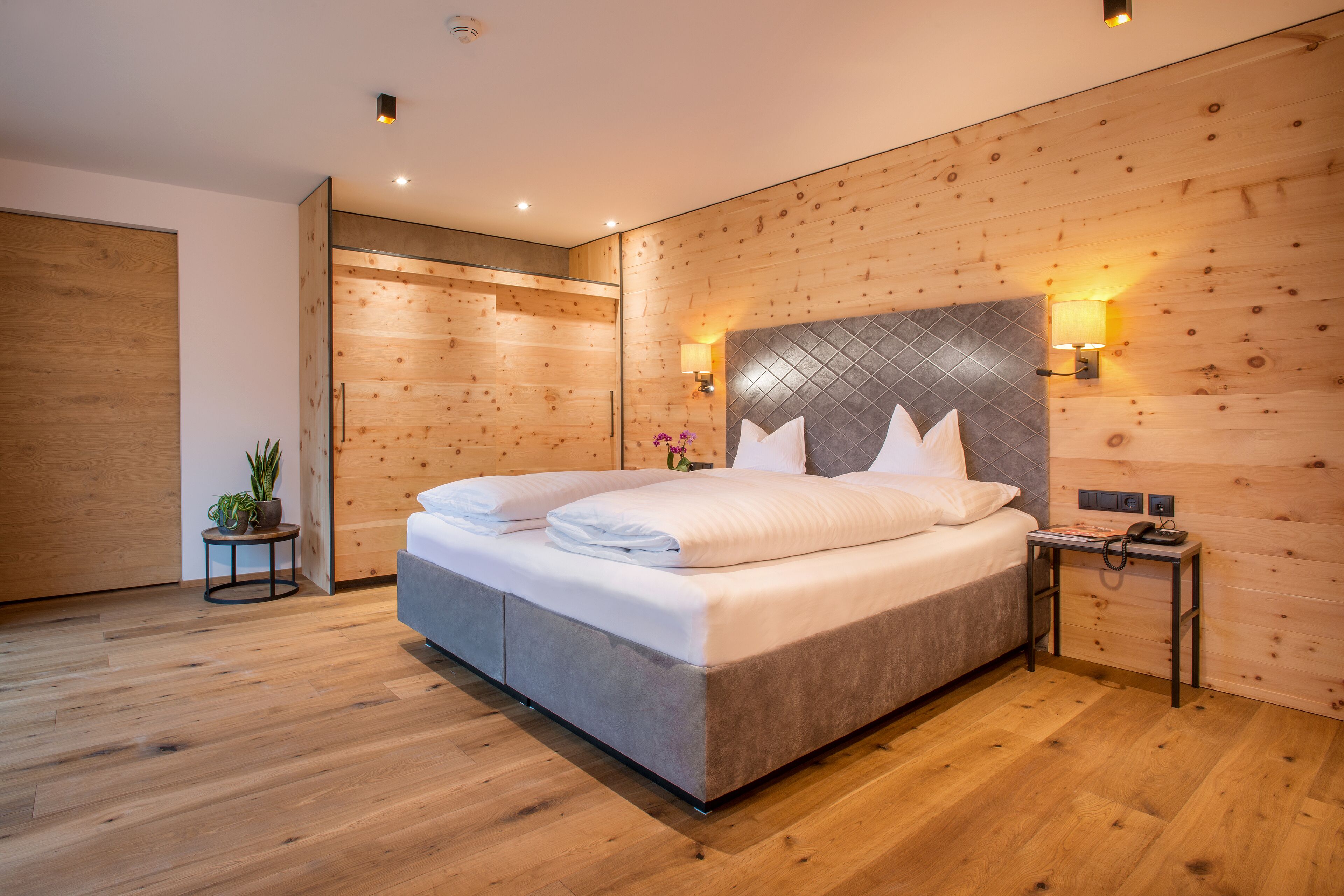 A modern bedroom with a large bed, wooden panel walls, and a small bedside table.