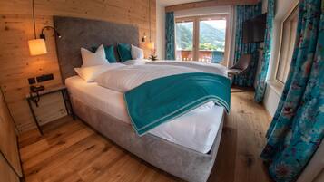 A large bed with a grey headboard, white bedding, and teal accents. There is a wooden floor, a nightstand with a lamp, and a chair by the window.
