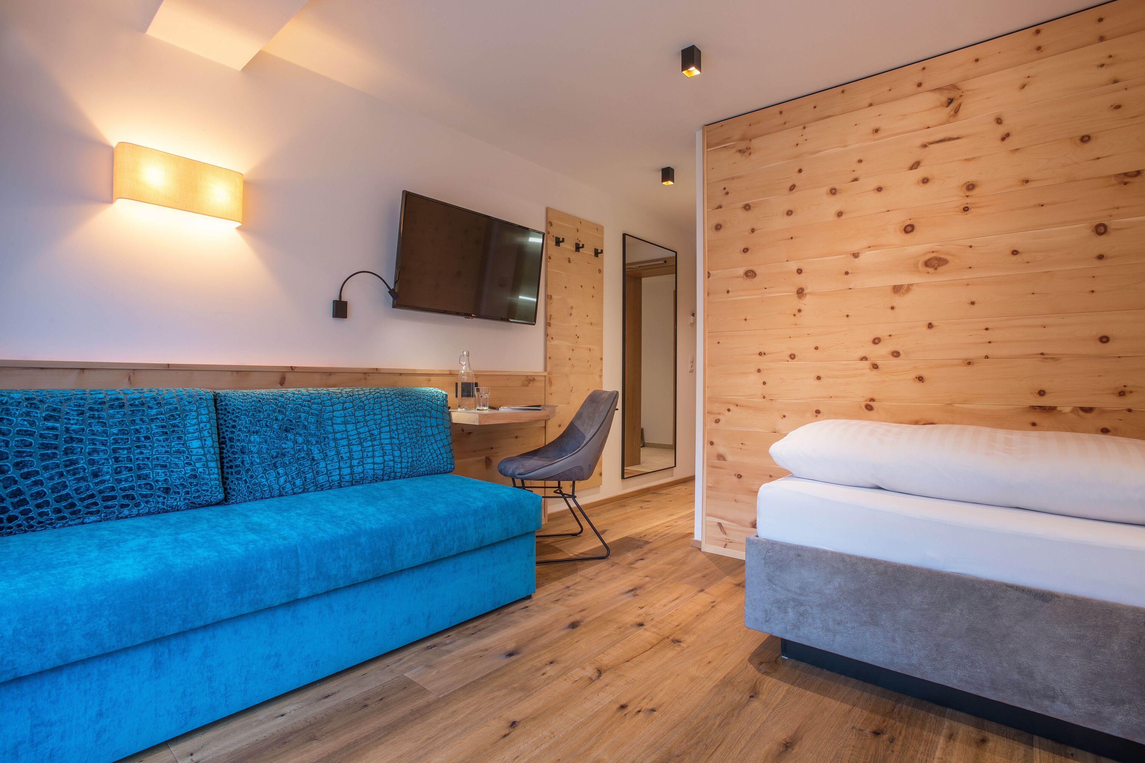 Una habitación moderna con un sofá azul, una silla gris, un revestimiento de madera y una cama con ropa blanca.