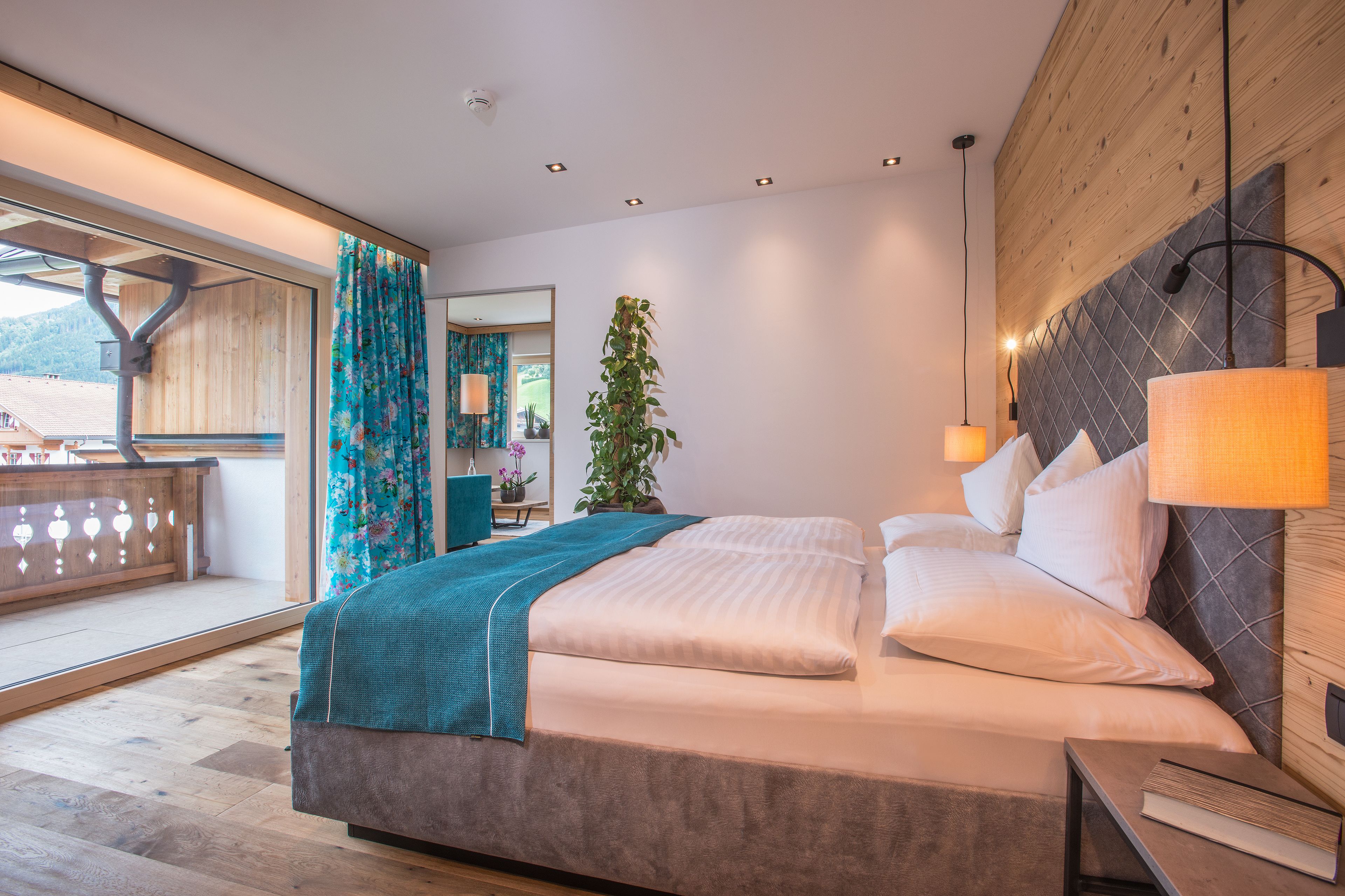 Habitación de hotel moderna con una cama grande, cabecera de madera, una planta en maceta y un balcón con vistas a las montañas.