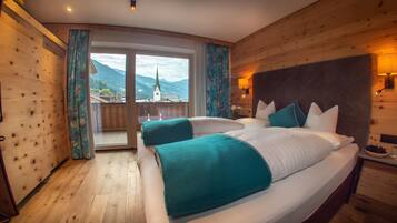 A double bed with teal and white bedding, wooden walls, a large window with curtains, and a view of mountains.
