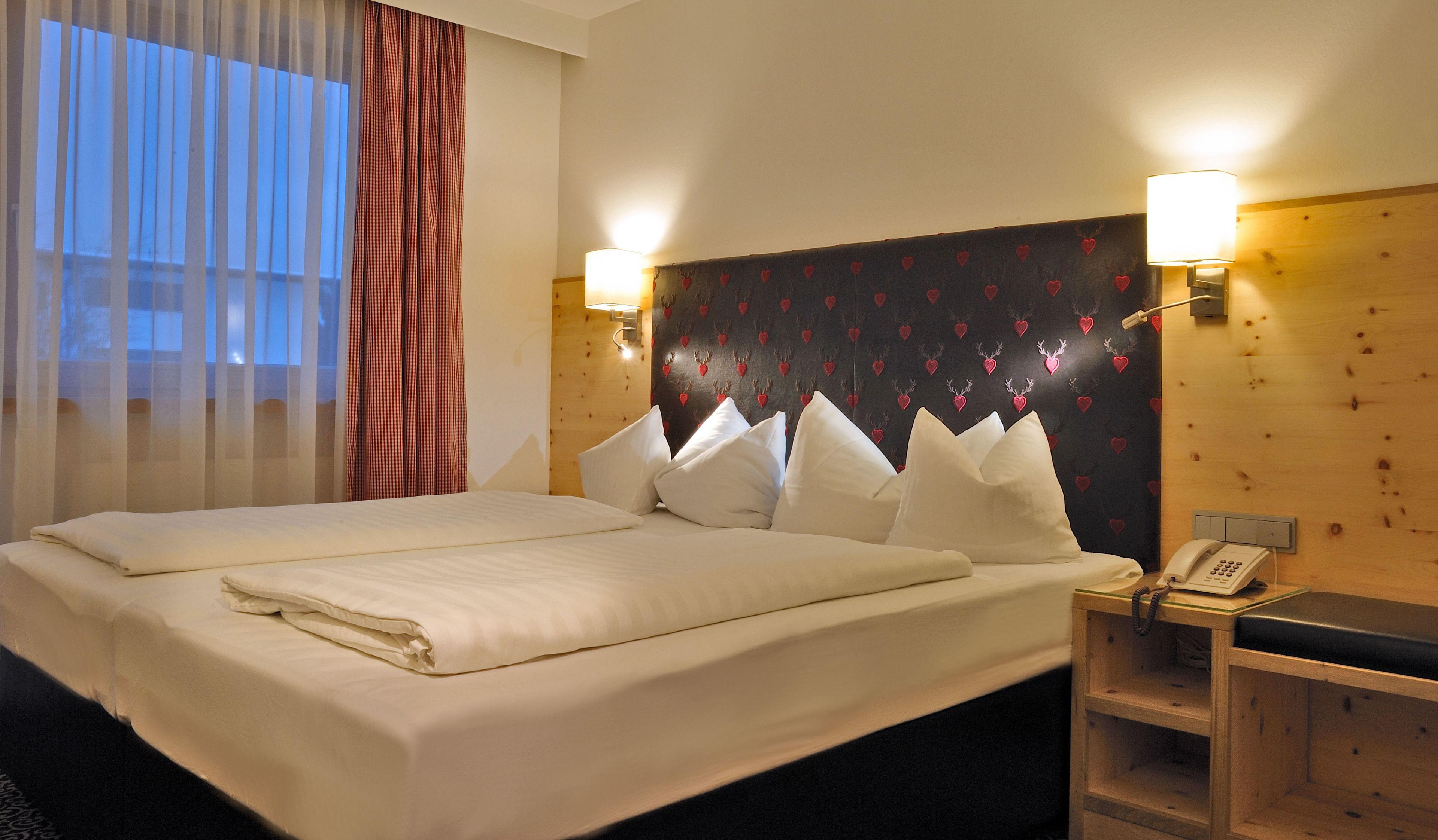 Habitación de hotel con una cama grande, dos lámparas de noche, un teléfono y un cabecero de madera con diseño de corazón.