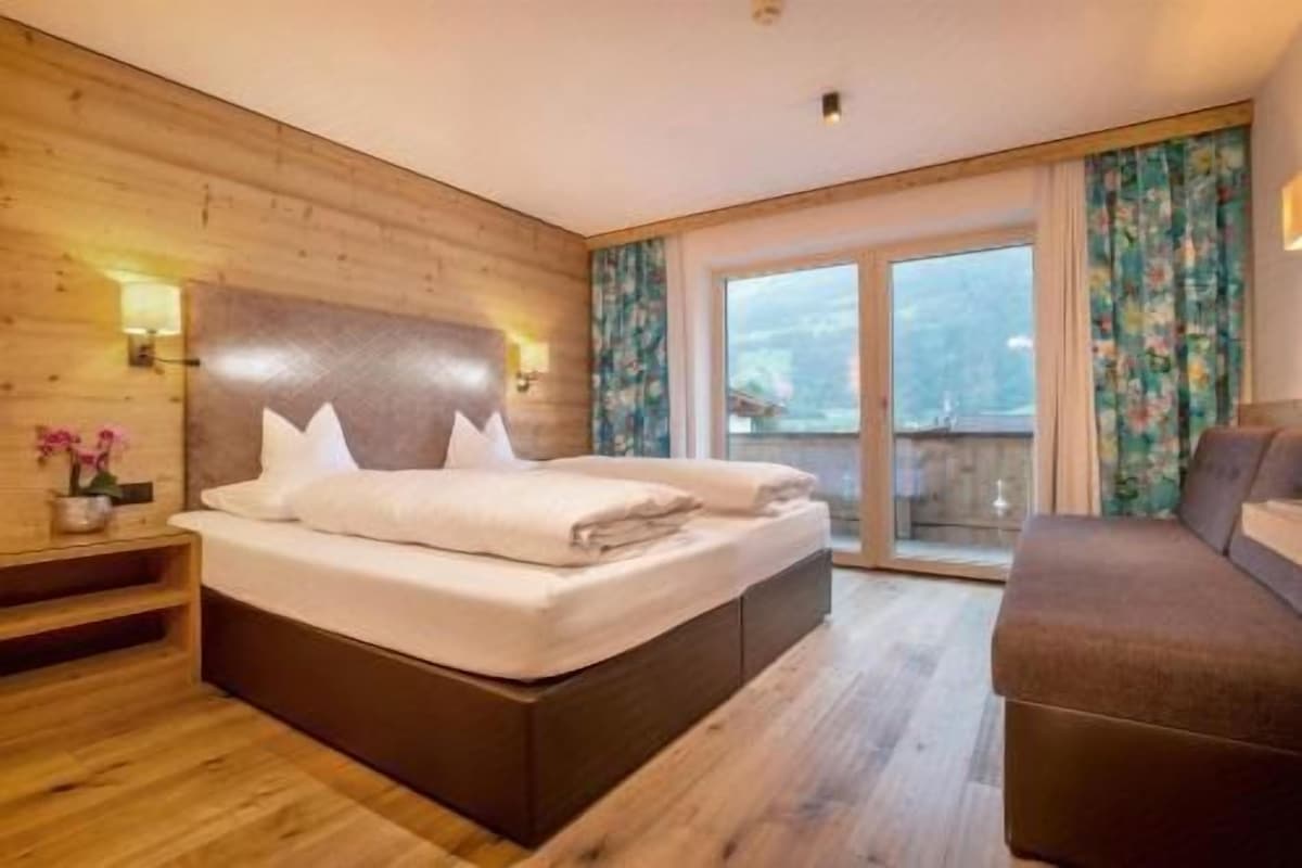 A bedroom with a large bed, a sofa, wooden walls, a balcony with a view, and a window with curtains.