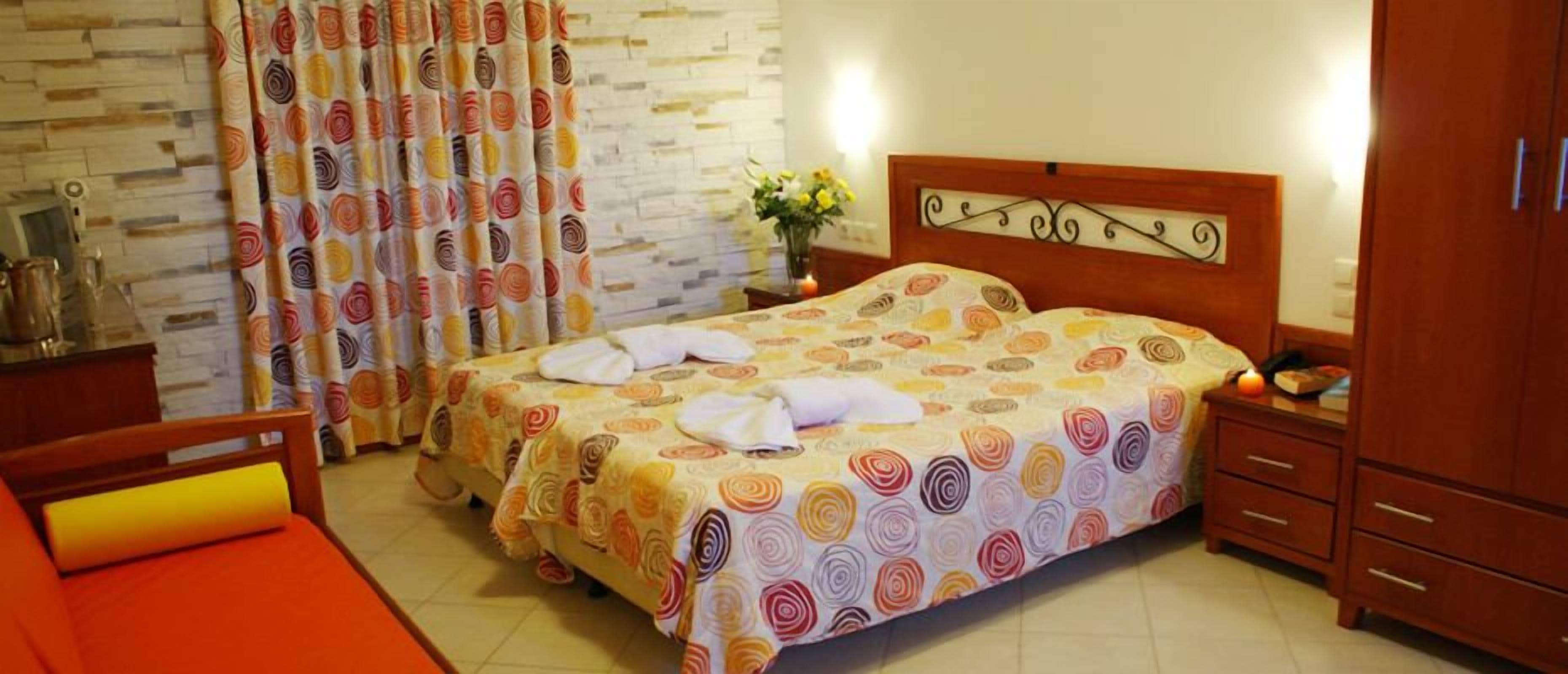Hotellihuone, jossa on sänky, puinen pääty, yöpöytä, työpöytä, tuoli ja kukkakuviollinen verho.