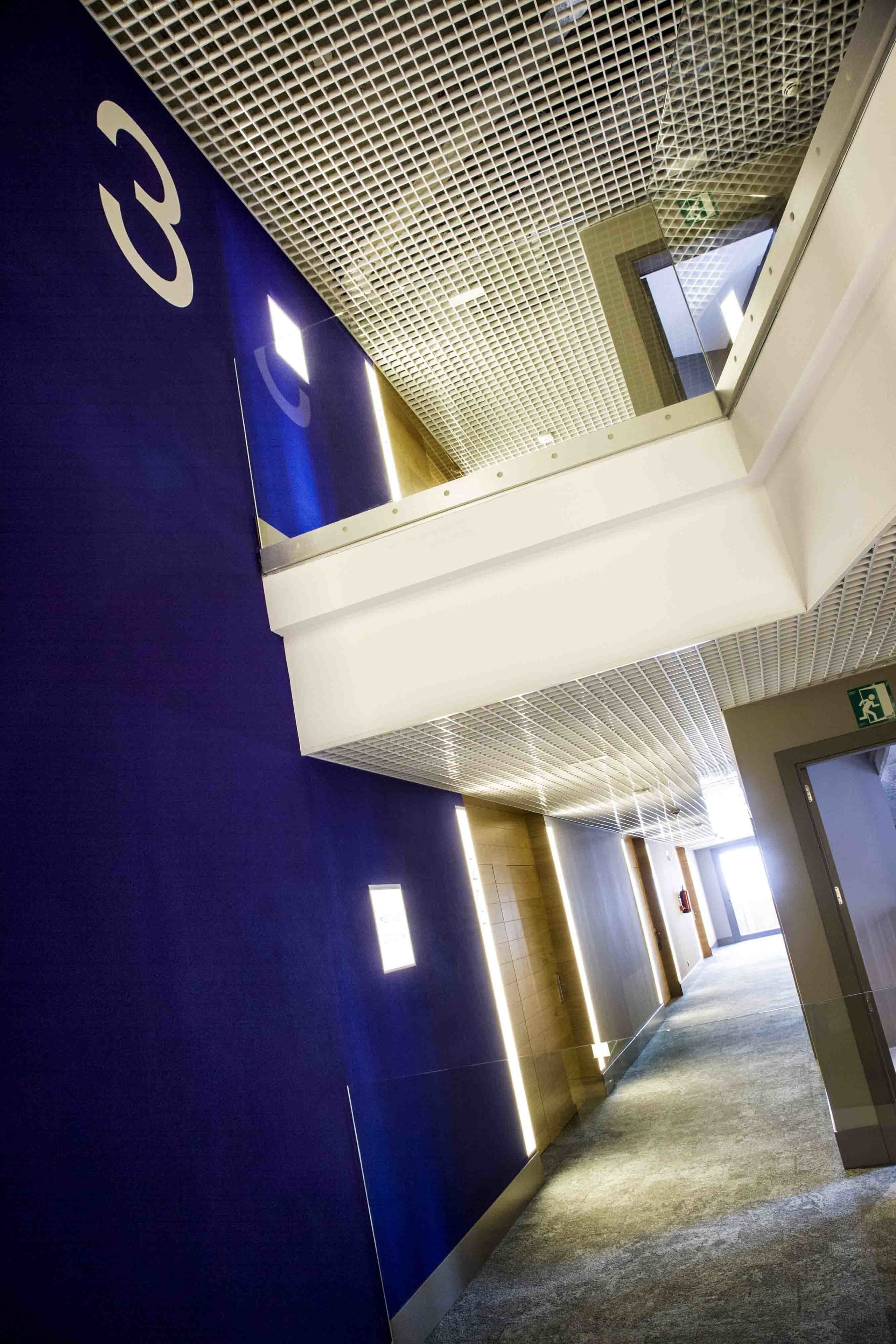 A corridor with a blue wall, a white ceiling with a grid pattern, and a visible emergency exit sign.