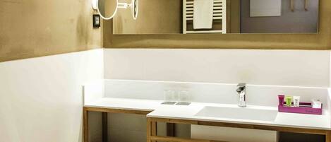 Un baño moderno con lavamanos, espejo y retrete blancos. Hay un estante de madera con toallas y un cesto de basura.