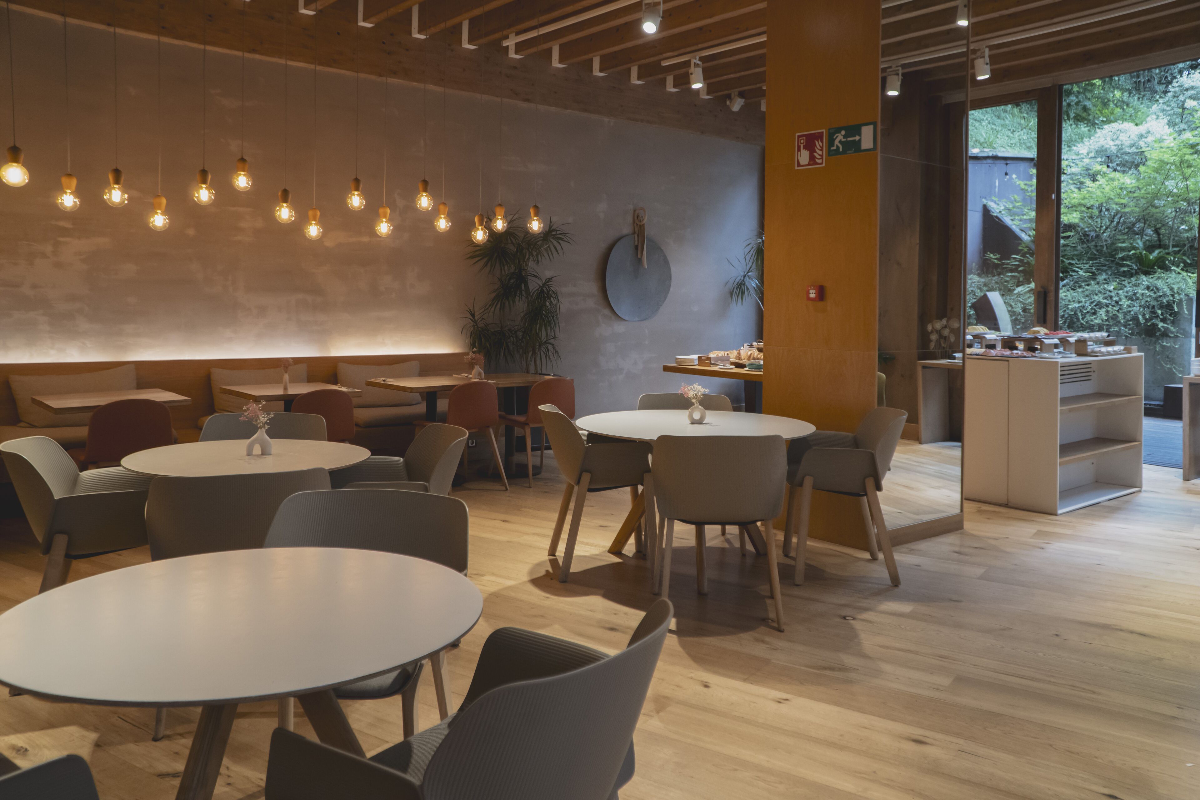 A modern cafe with round tables and chairs, hanging light fixtures, and a view of greenery outside.