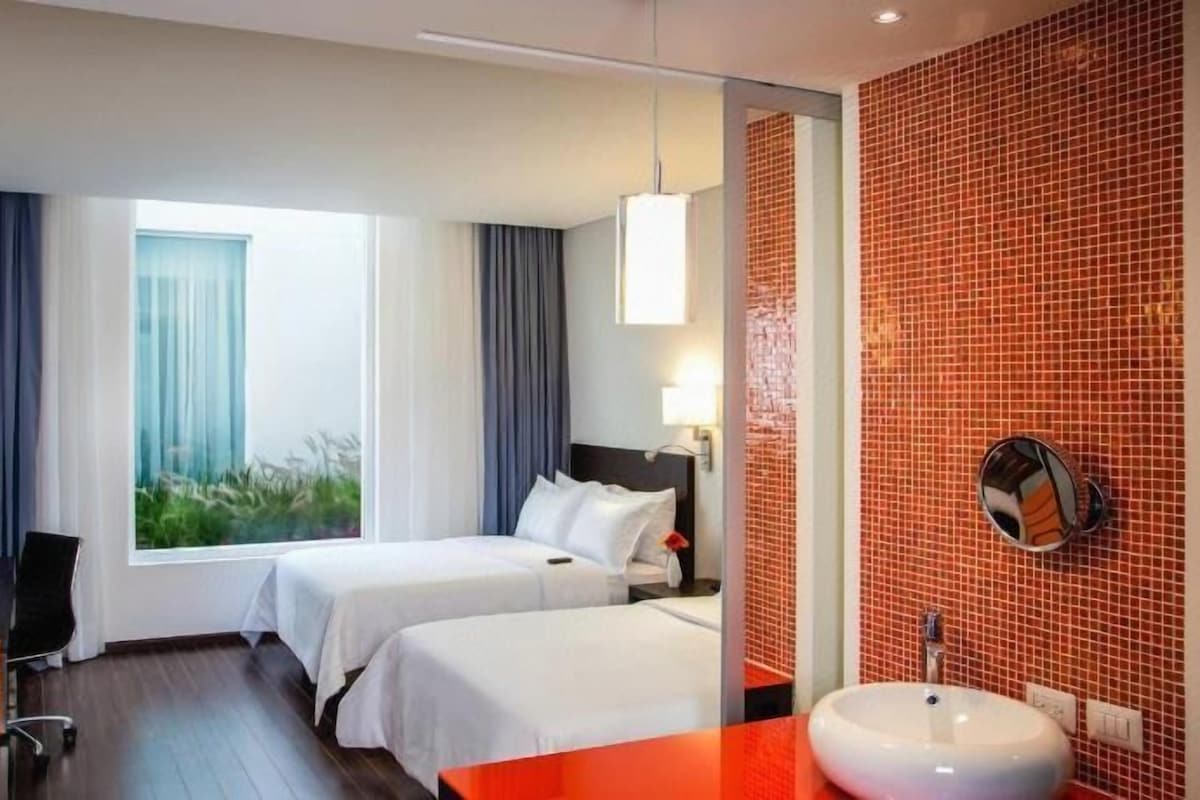 Habitación de hotel con dos camas, un escritorio, una silla, un televisor, un ventanal con cortinas y un baño con lavamanos y espejo.