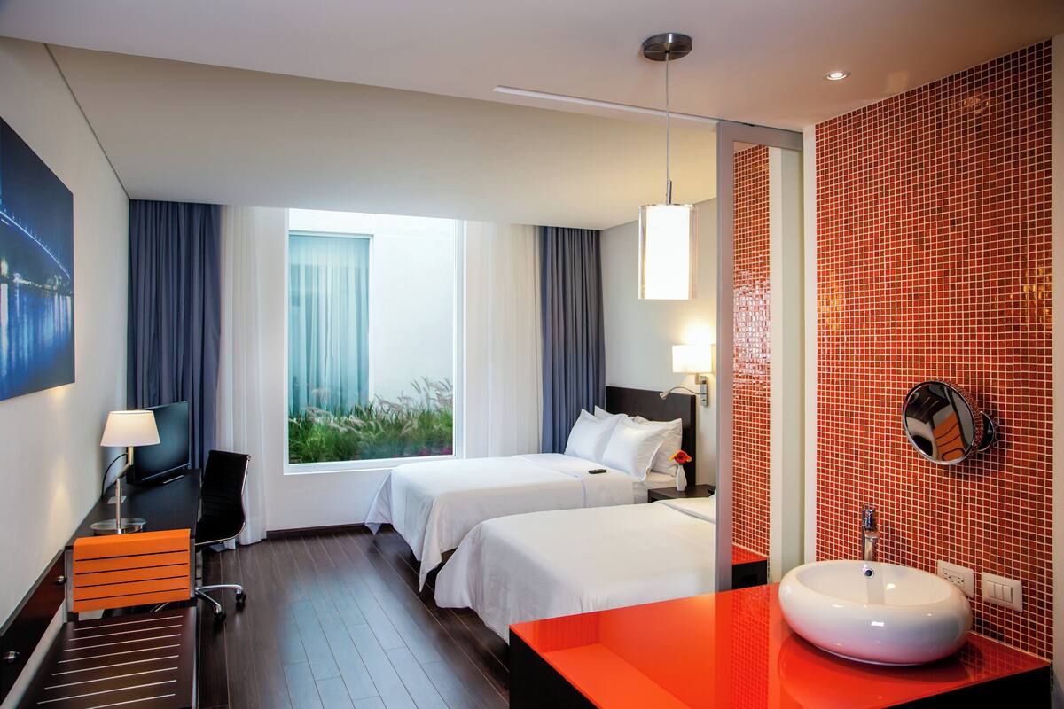 A hotel room with two beds, a desk, a chair, a TV, a window with curtains, a red tiled wall, and a round mirror.