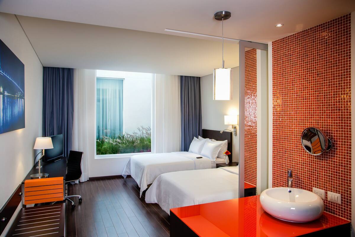 A hotel room with two beds, a desk, a chair, a large window, a bathroom with a red tiled wall, and a round mirror.