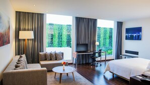 A modern hotel room with a sofa, a small round table with a vase, a bed, a desk with a TV, and large windows with curtains. - Fiesta Inn Plaza Central Aeropuerto (Mexico City)