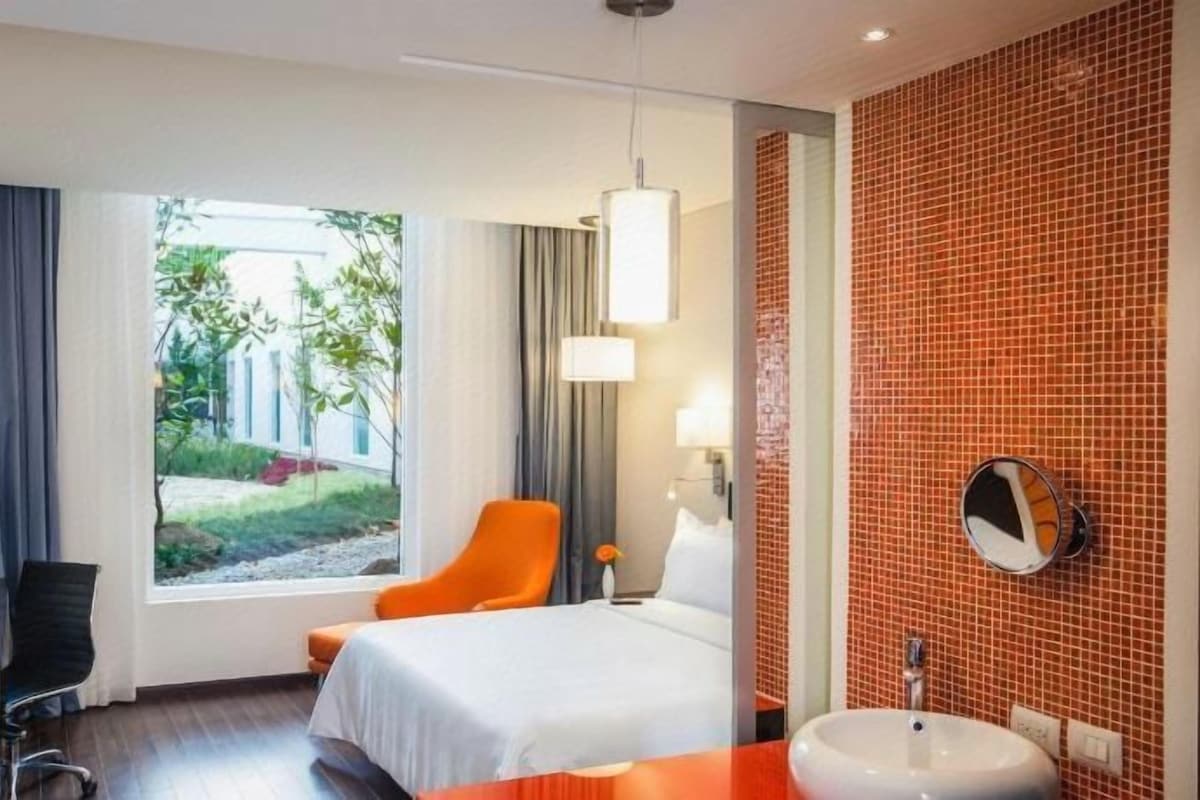 Habitación de hotel con cama, escritorio, silla, baño con lavamanos y vista a un jardín.