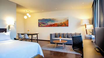 A hotel room with a bed, a sofa, a small table with a vase, a TV, and a painting on the wall.
