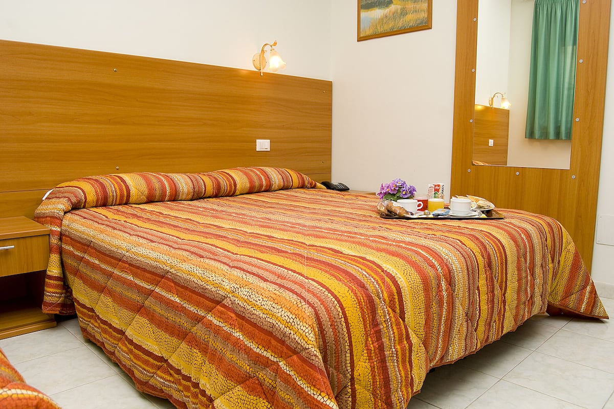 Un lit bien fait, recouvert d’une couvre-lit à rayures, une table de chevet avec une lampe, et une petite table sur laquelle se trouve un plateau avec divers objets.