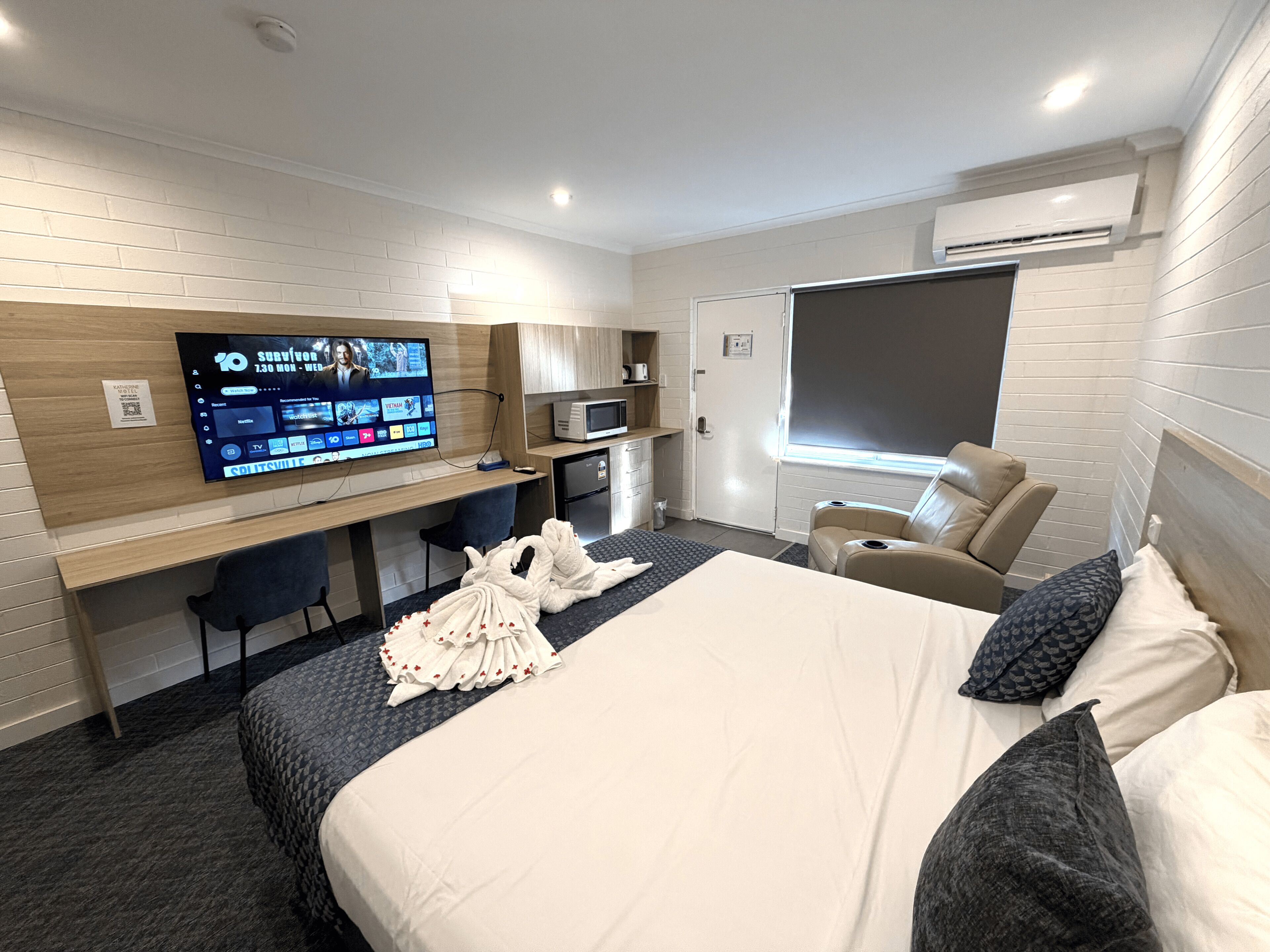 A hotel room with a bed, a desk with a computer, a TV, a microwave, and a massage chair.