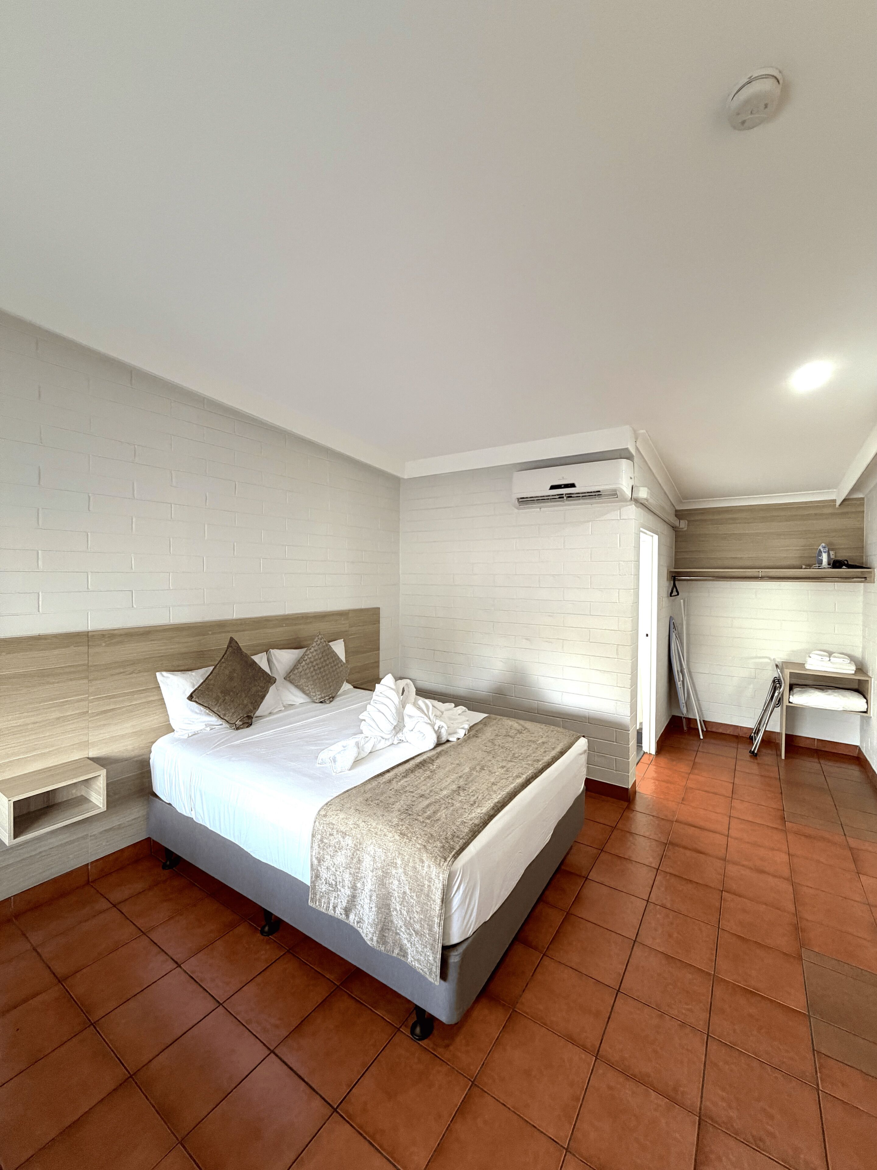 A hotel room with a bed, a wooden headboard, a white brick wall, an air conditioning unit, and a small seating area with a table and chairs.