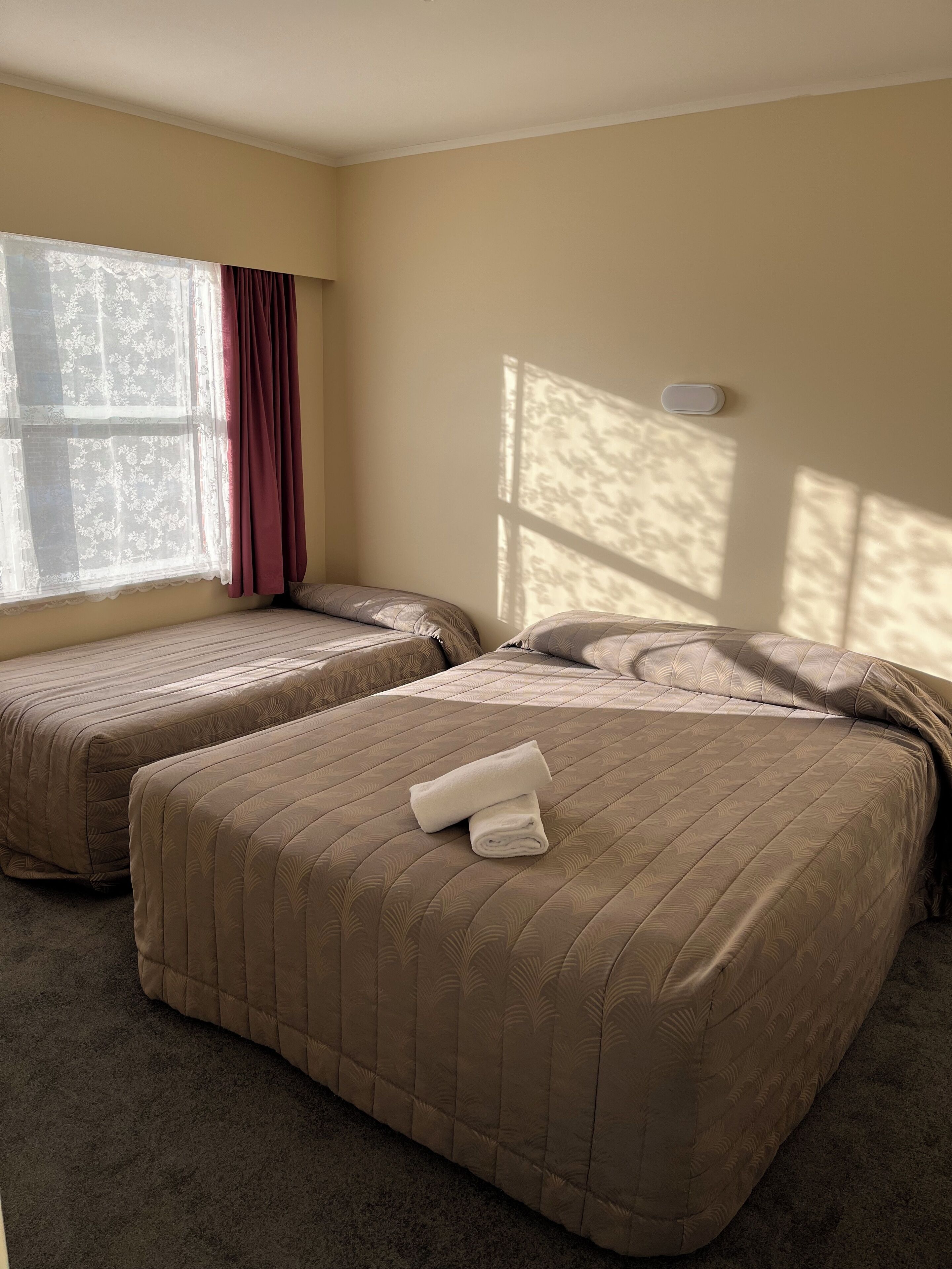 A hotel room with two beds, a window with curtains, and a thermostat on the wall.