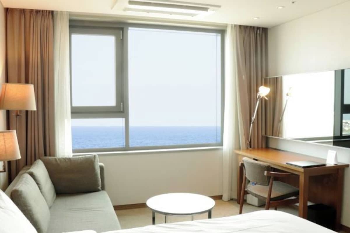 A hotel room with a bed, a desk, a chair, a small table, and a large window with a view of the sea.
