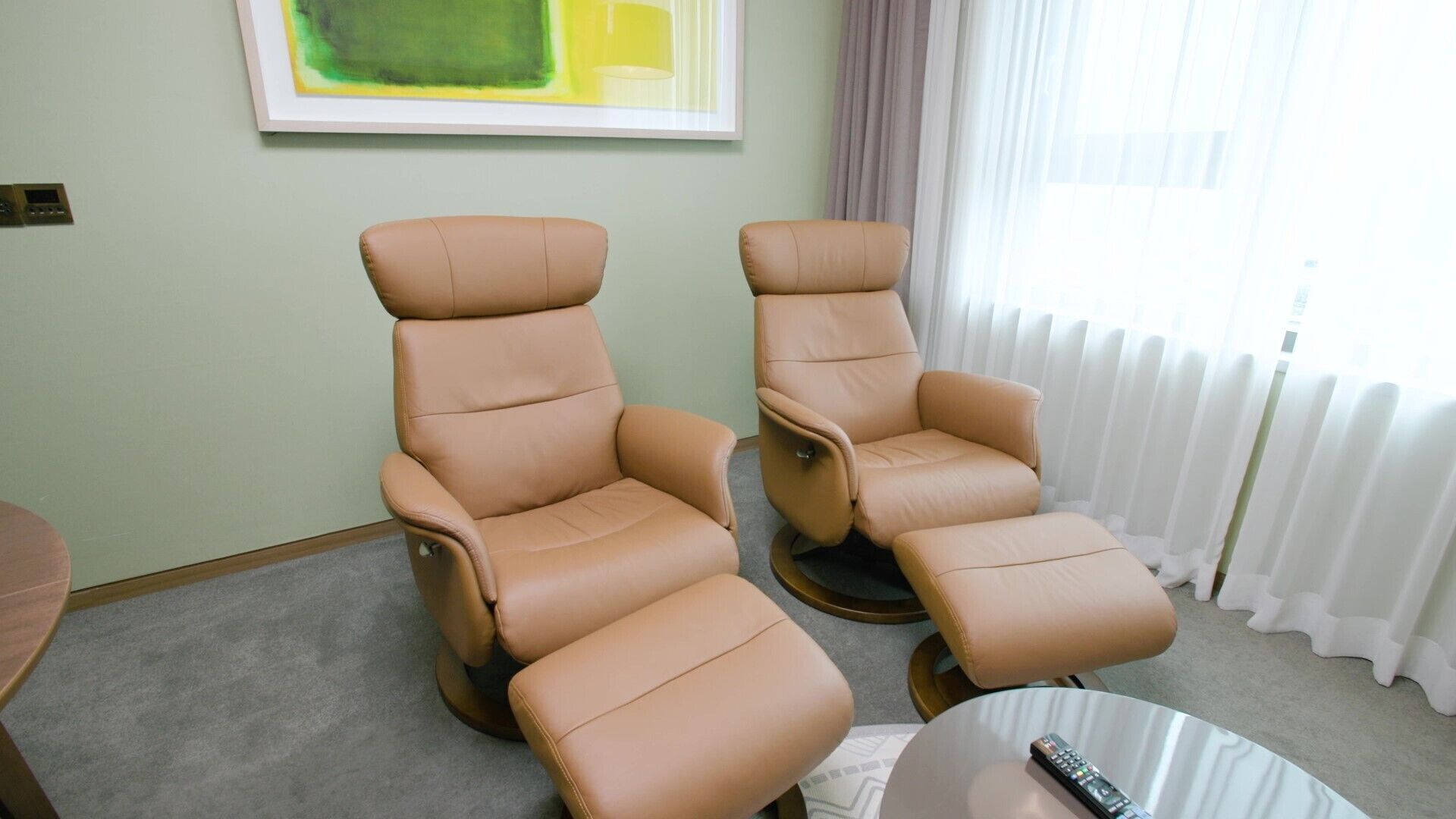 A room with two reclining massage chairs, a small round table with a remote control, and a painting on the wall.